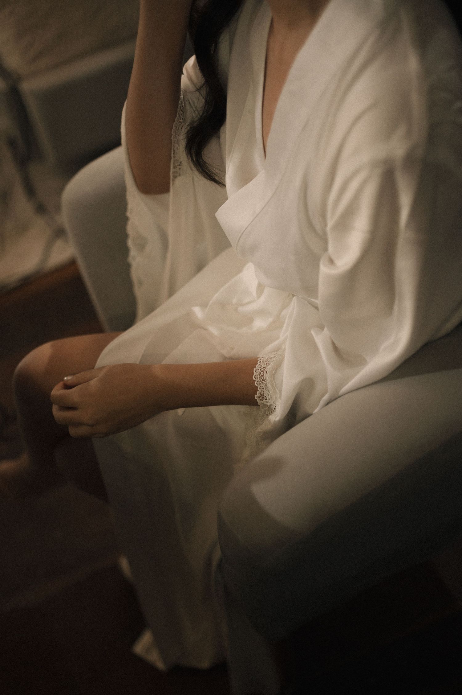 Woman in white satin robe with lace trim sits gracefully on a chair, perfect bridal getting ready attire.
