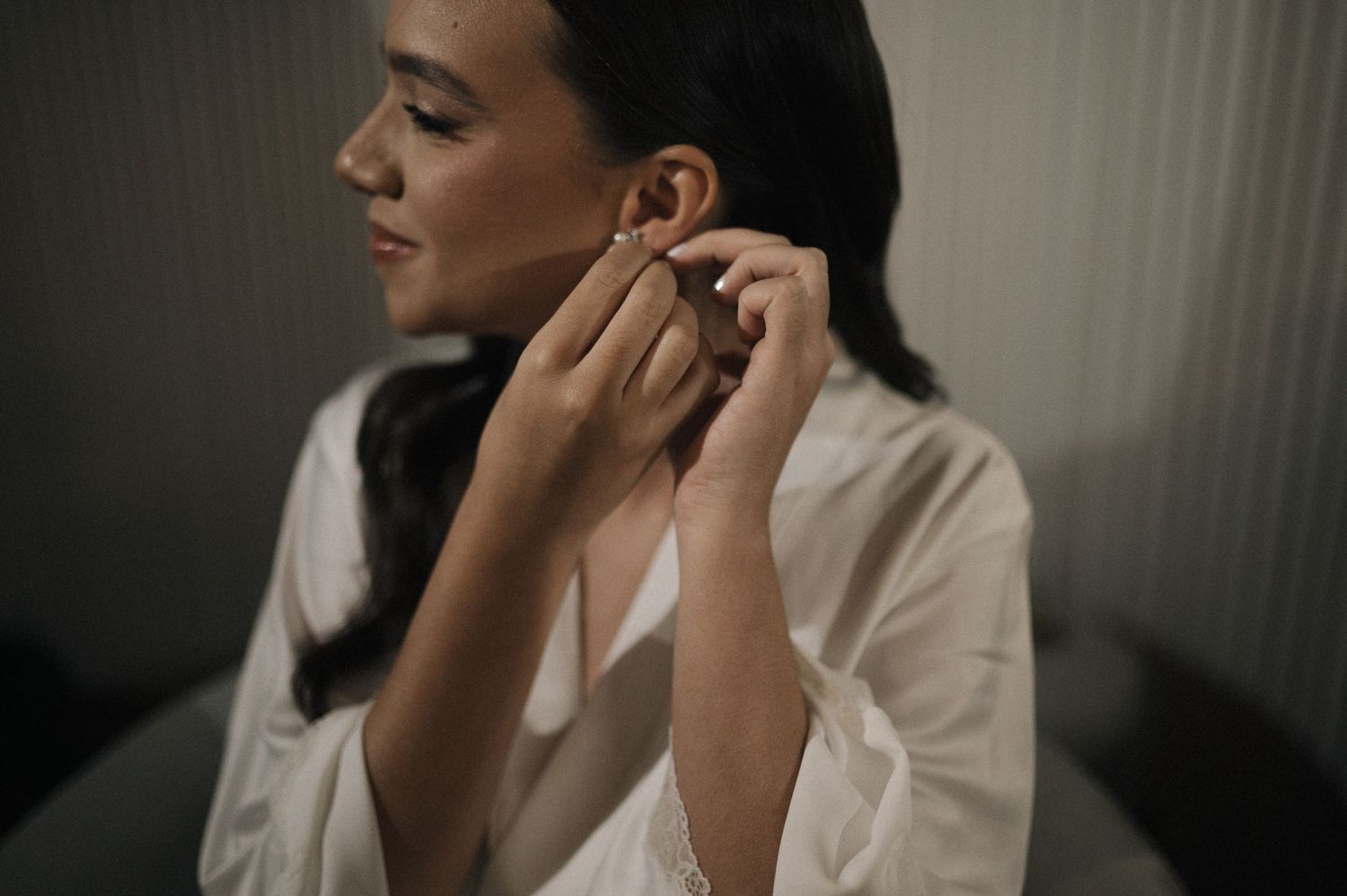 Woman in white robe putting on earrings, elegant bridal getting ready moment.