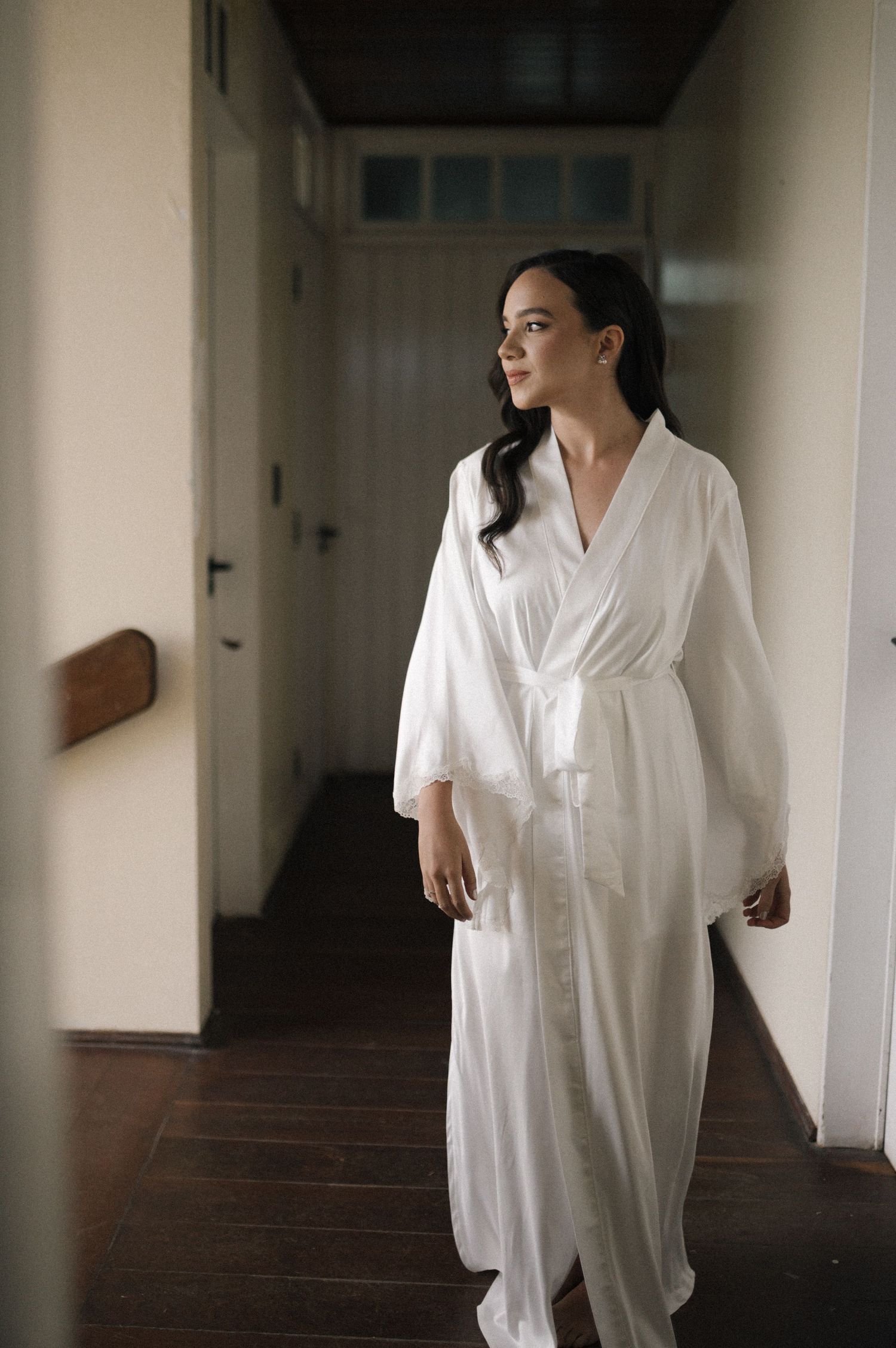 Woman in white satin robe standing in a hallway, gazing to the side with elegant poise.