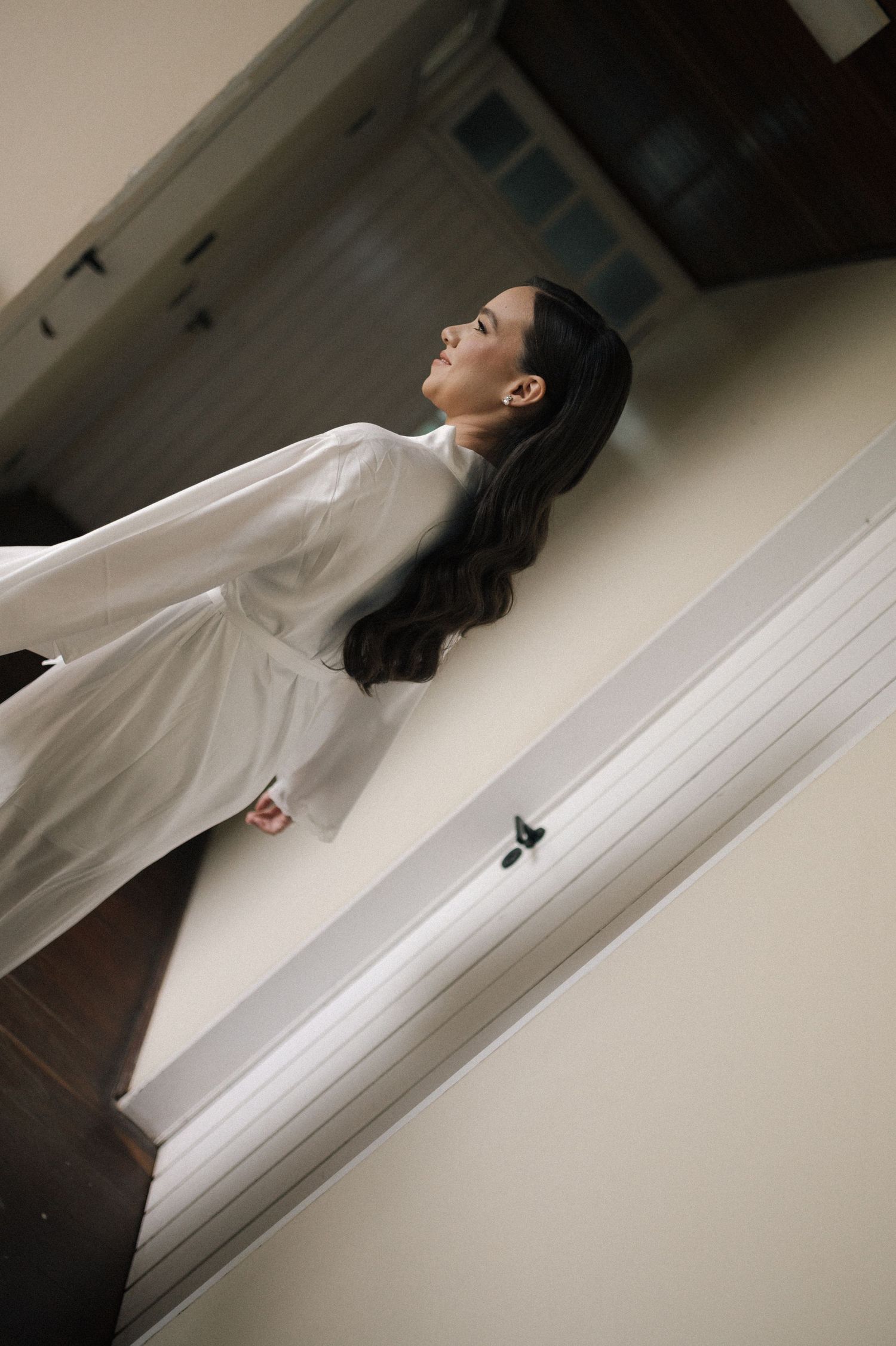 Woman in white dress gazing upward while standing near staircase railing in elegant interior.