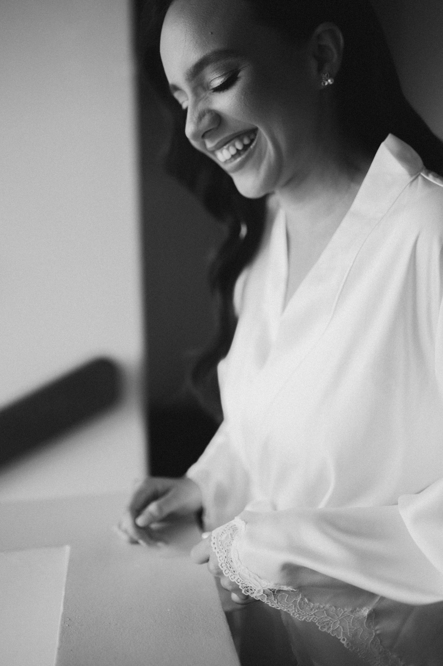 Black and white photo of a smiling bride in a white lace robe reading a letter on her wedding day.