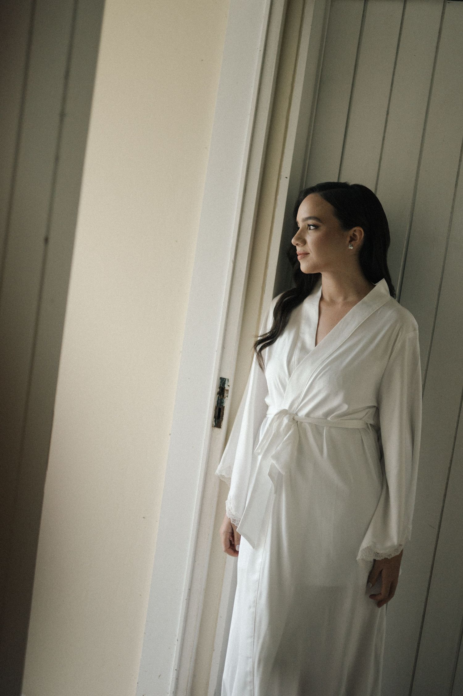 Woman in white silk robe standing by a door, gazing thoughtfully to the side in soft natural light.