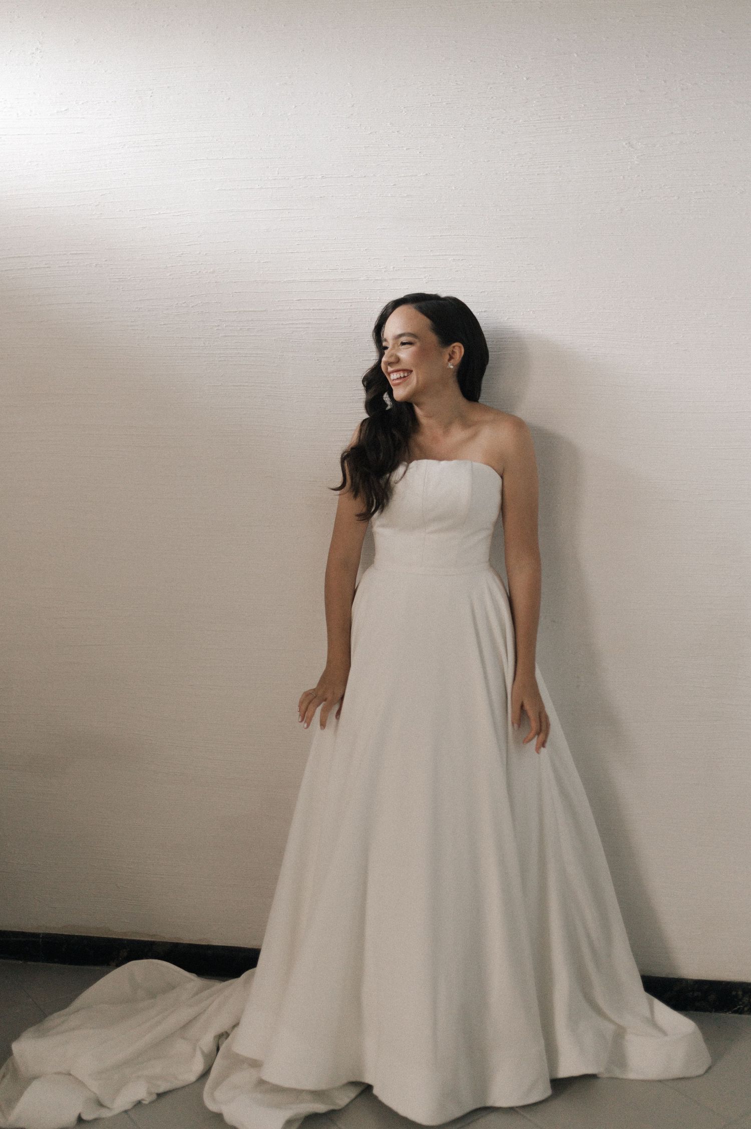 Smiling bride in a strapless white A-line wedding gown with a train, posing against a light wall.