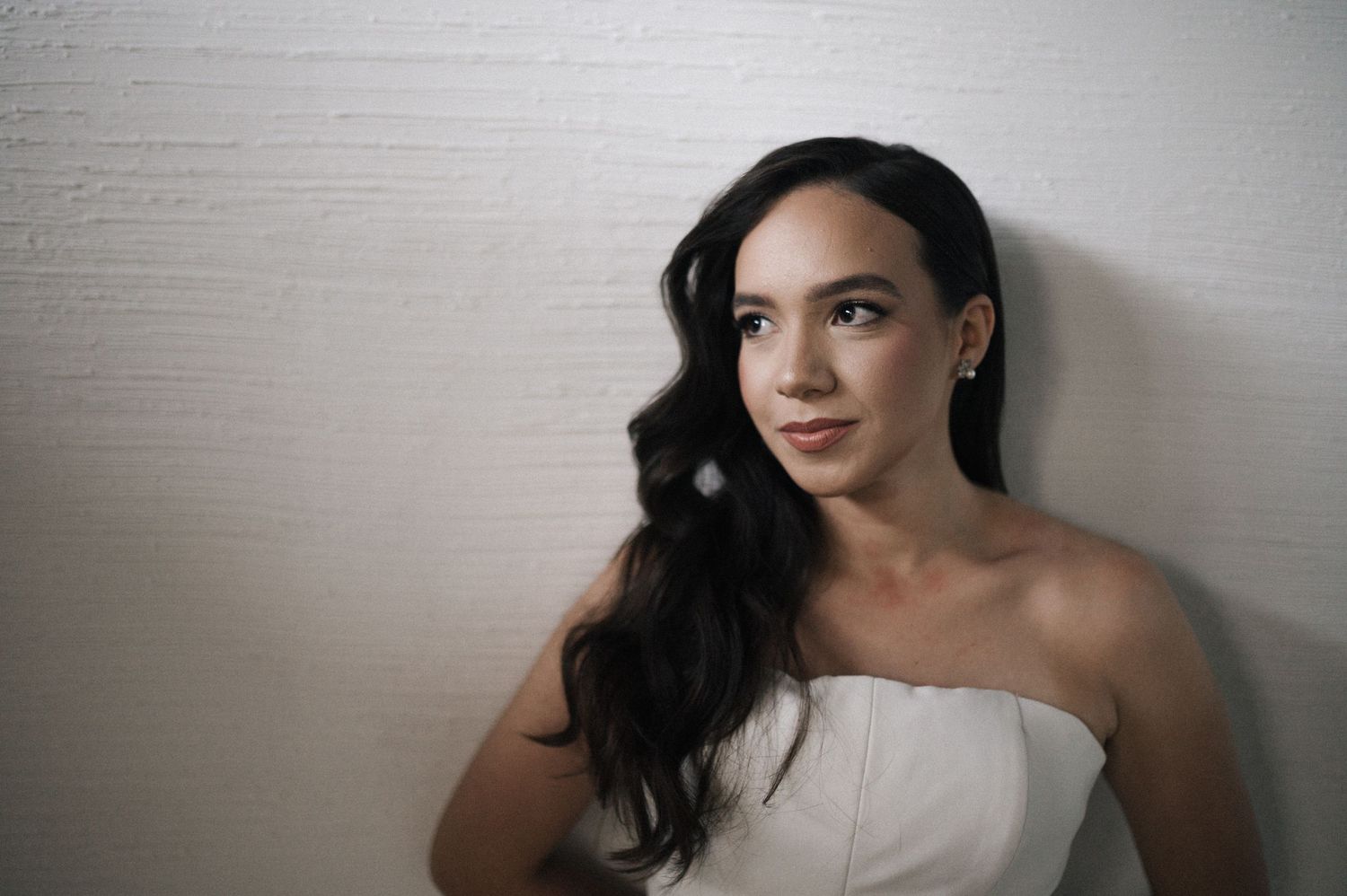 Young woman in white strapless dress with dark wavy hair poses against a light wall, looking away thoughtfully.