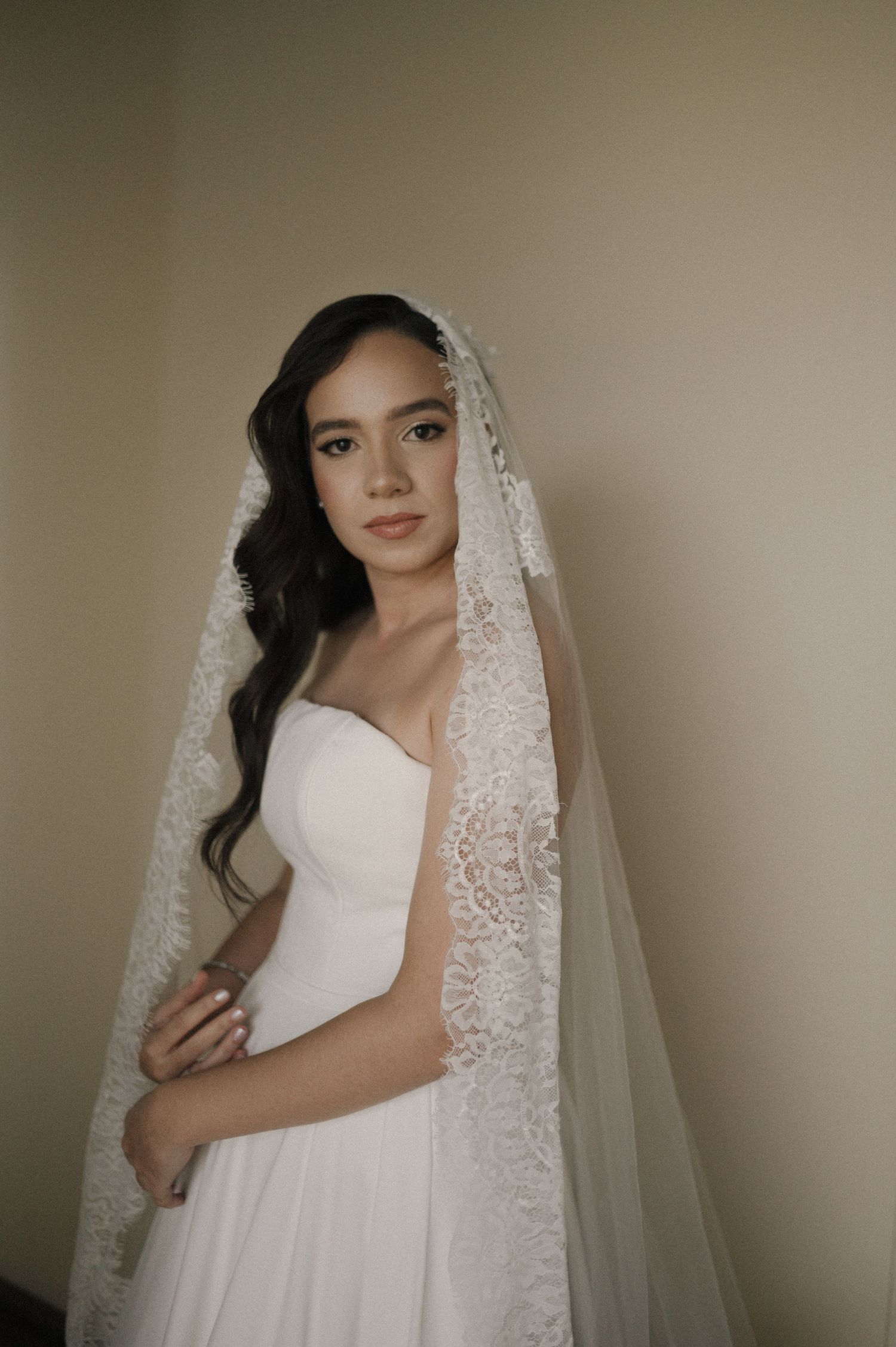 A bride in a white strapless gown and lace veil poses elegantly with arms crossed in a softly lit room.