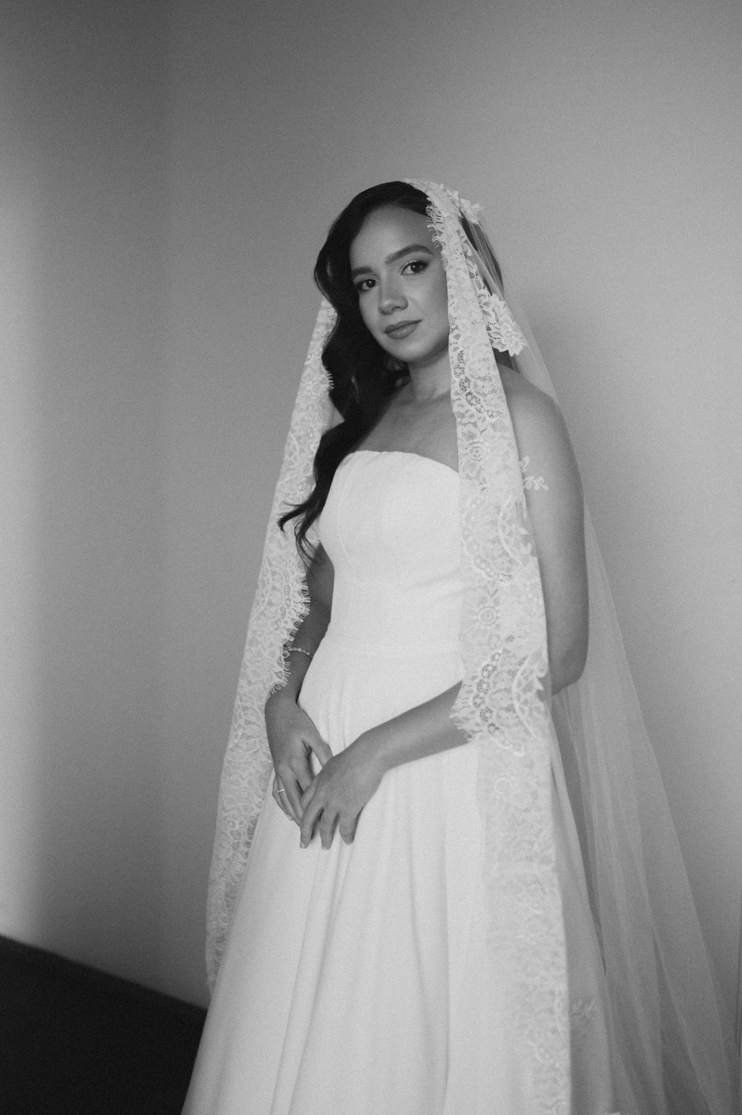 Black and white portrait of a bride in a strapless gown with lace-trimmed cathedral veil, posing elegantly.