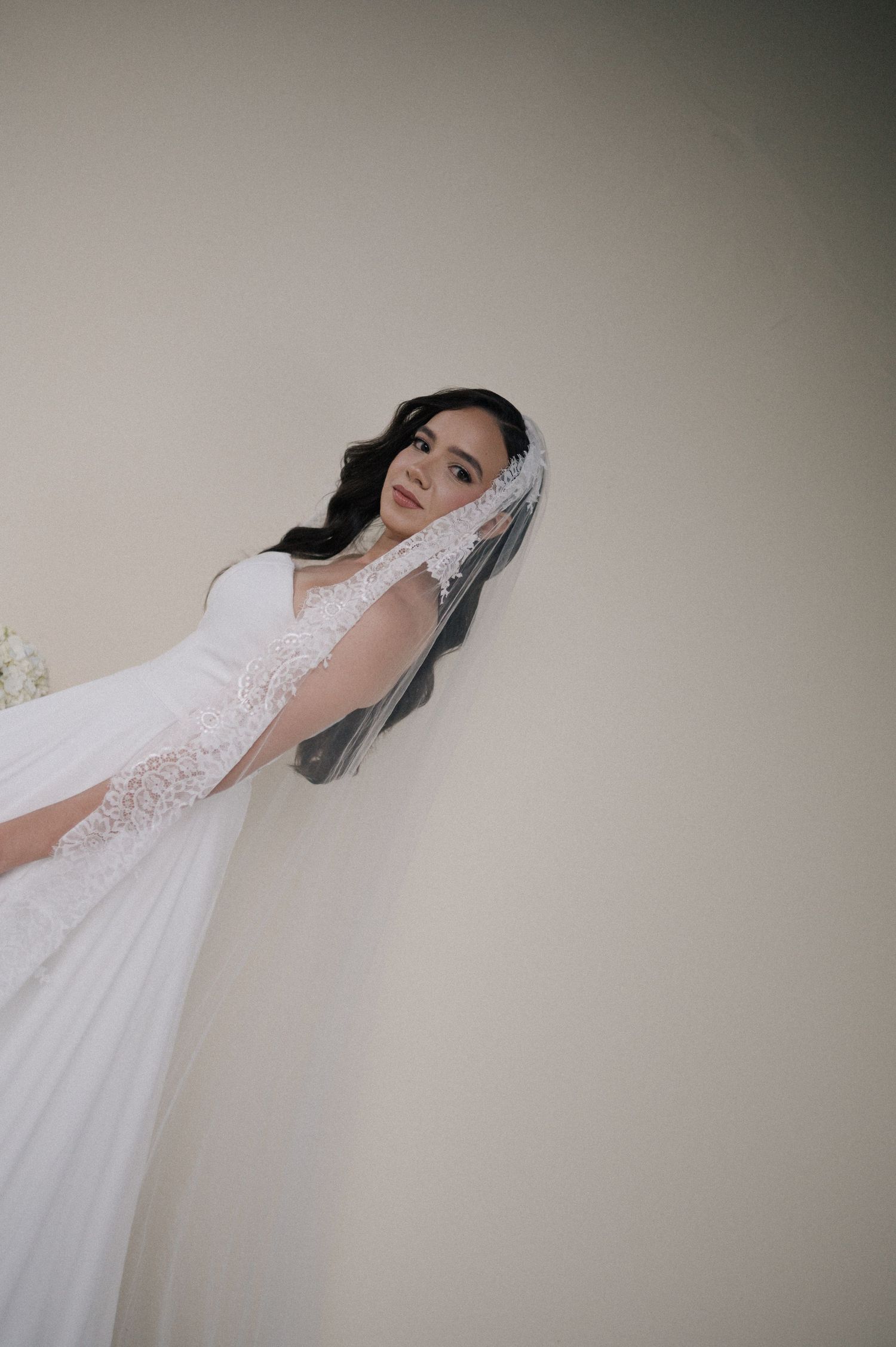 Bride in a white lace wedding gown and long veil poses gracefully against a neutral wall.