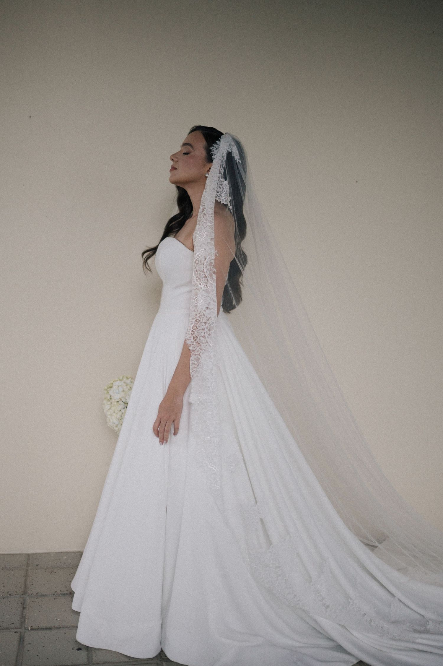 Elegant bride in a white ball gown with lace veil holding a bouquet, posing gracefully against a neutral wall.