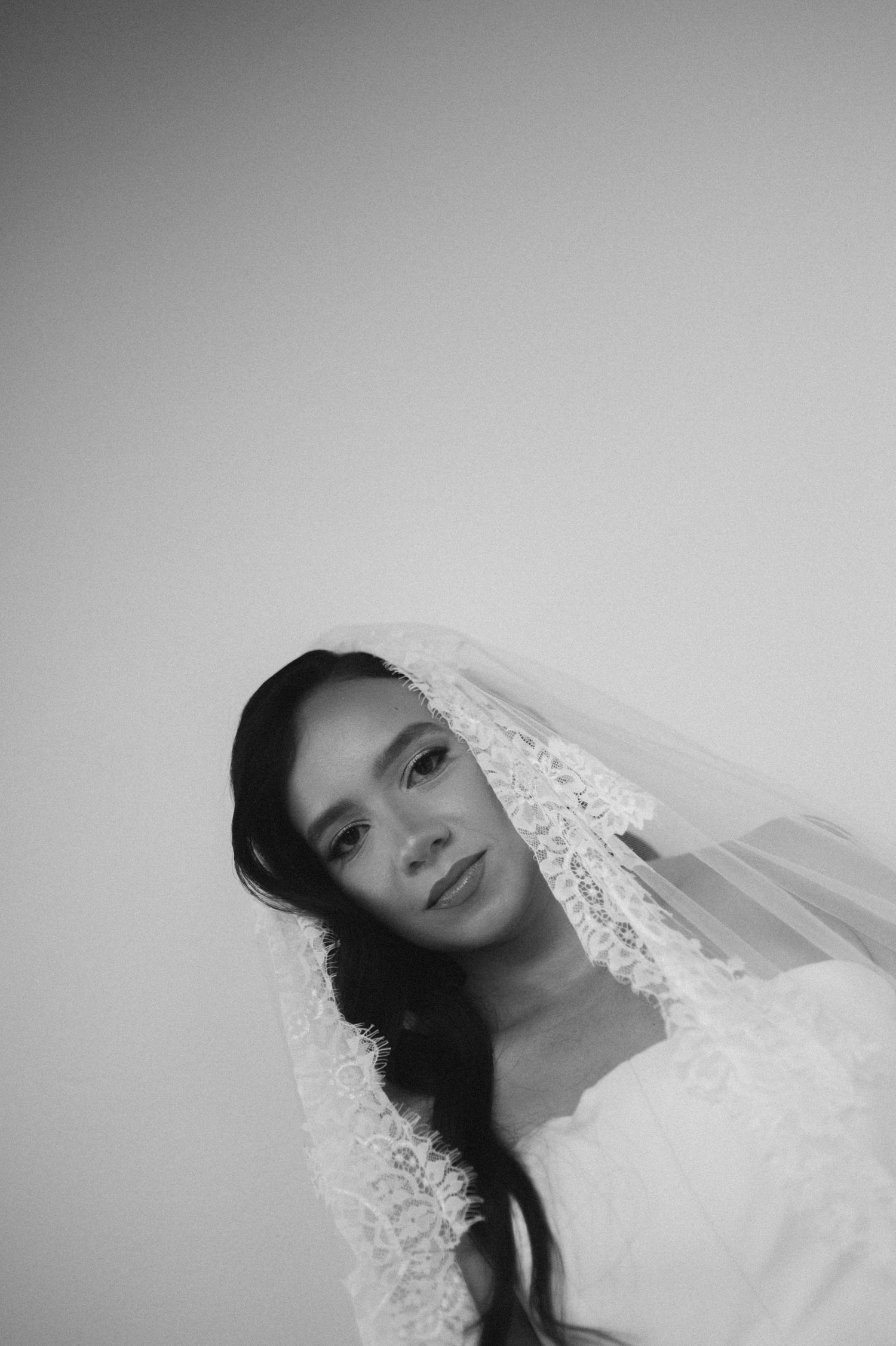 Black and white portrait of a bride wearing a lace veil, gazing softly at the camera against a light background.