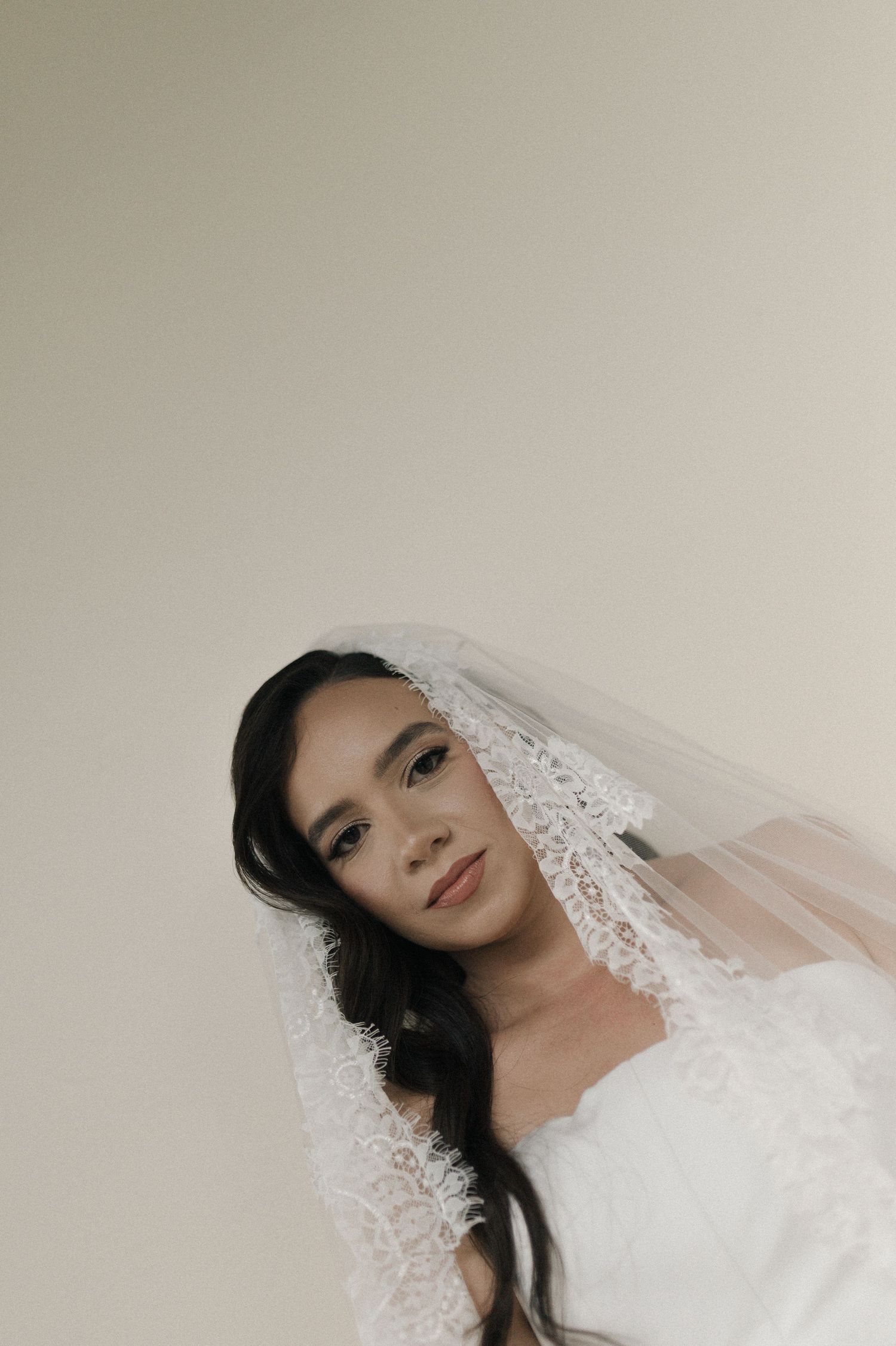 Bride wearing a lace-trimmed veil and white gown, posing elegantly against a neutral background.