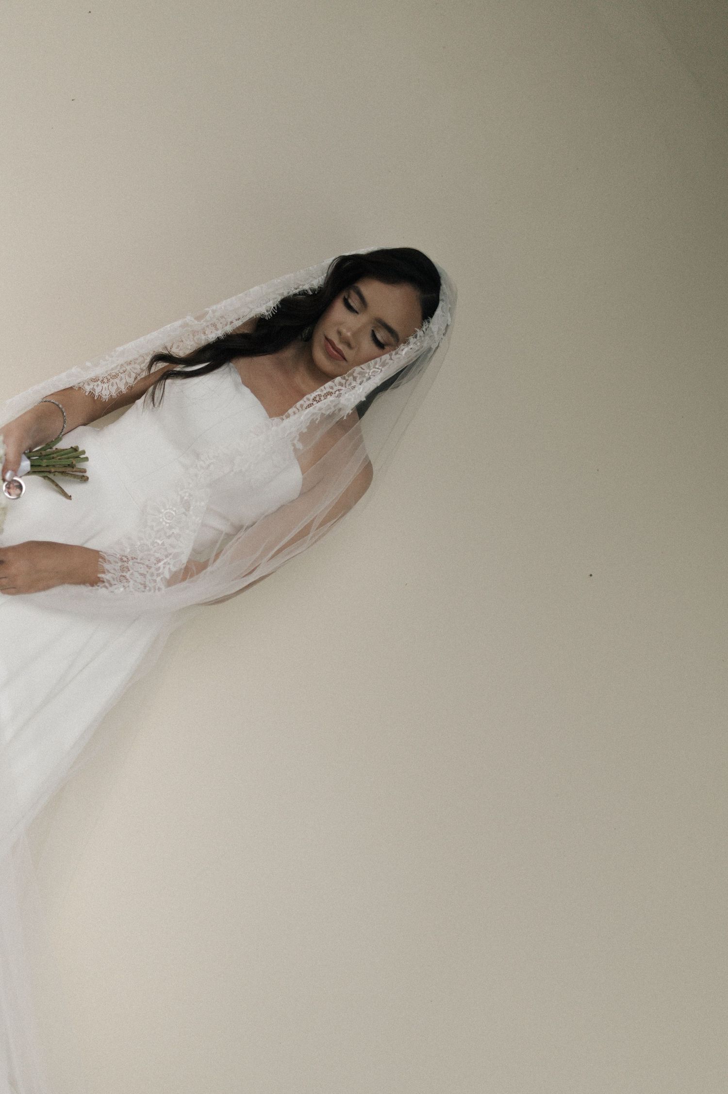 A bride in a flowing white gown and lace veil stands gracefully with eyes closed, holding a bouquet.