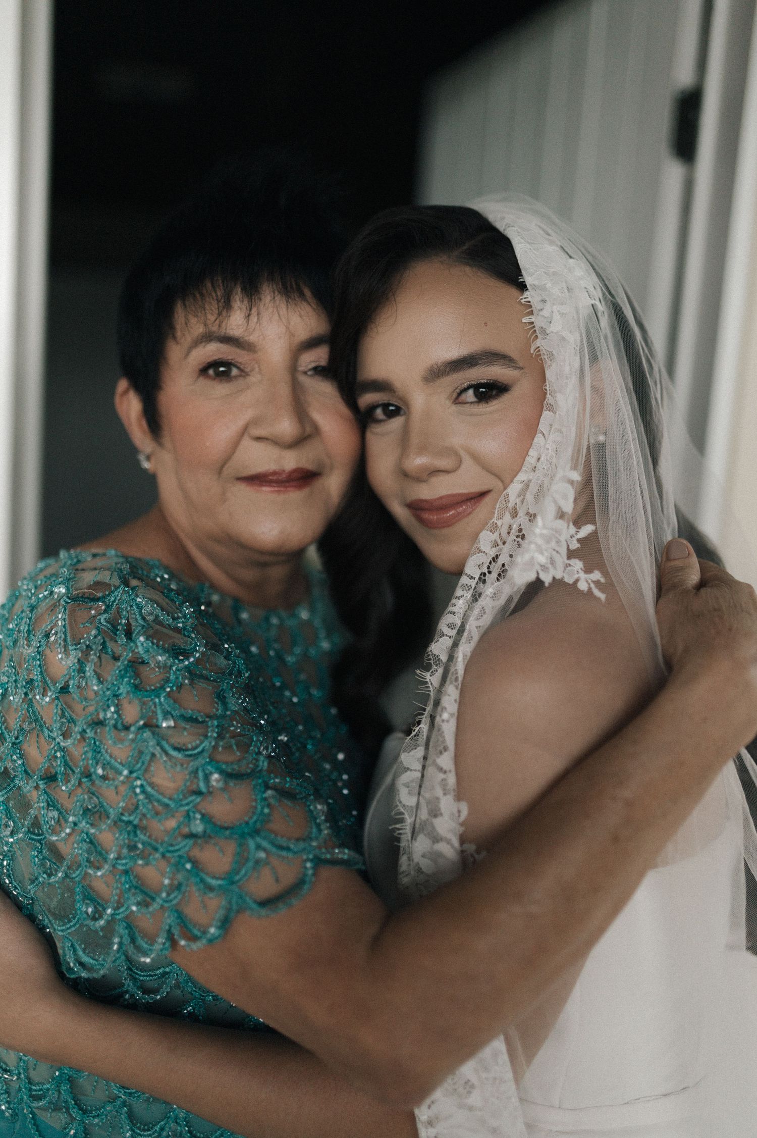 A bride in a white lace veil embraces an older woman in a teal beaded gown, sharing a warm moment.