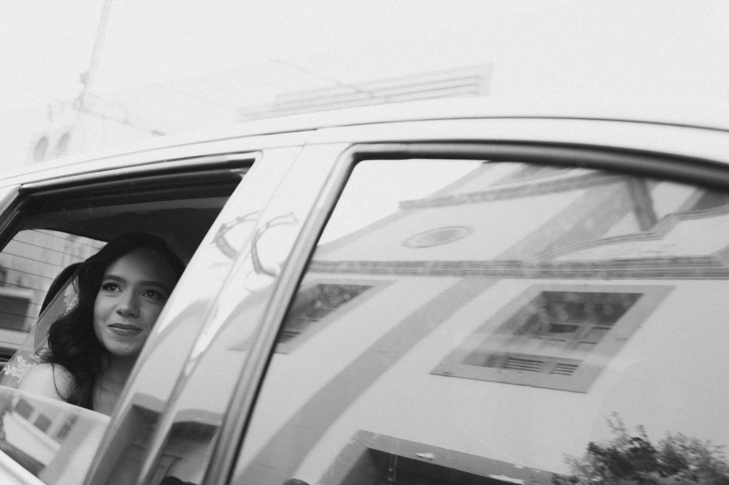 Black and white photo of a woman in a hat gazing out of a car window with building reflections.