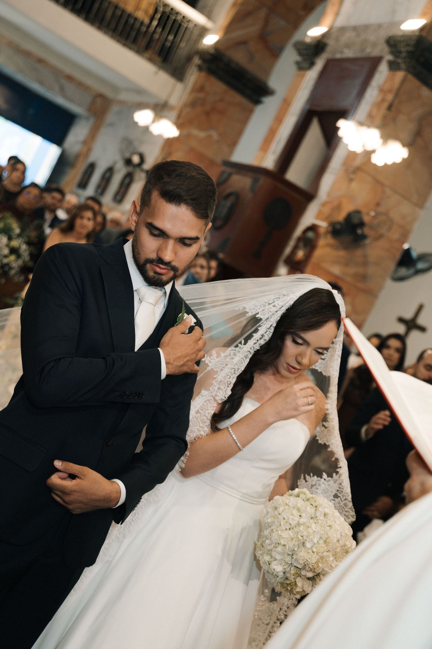Bride and groom share a tender moment at church altar, bride in white gown with veil holding bouquet, groom in black suit.
