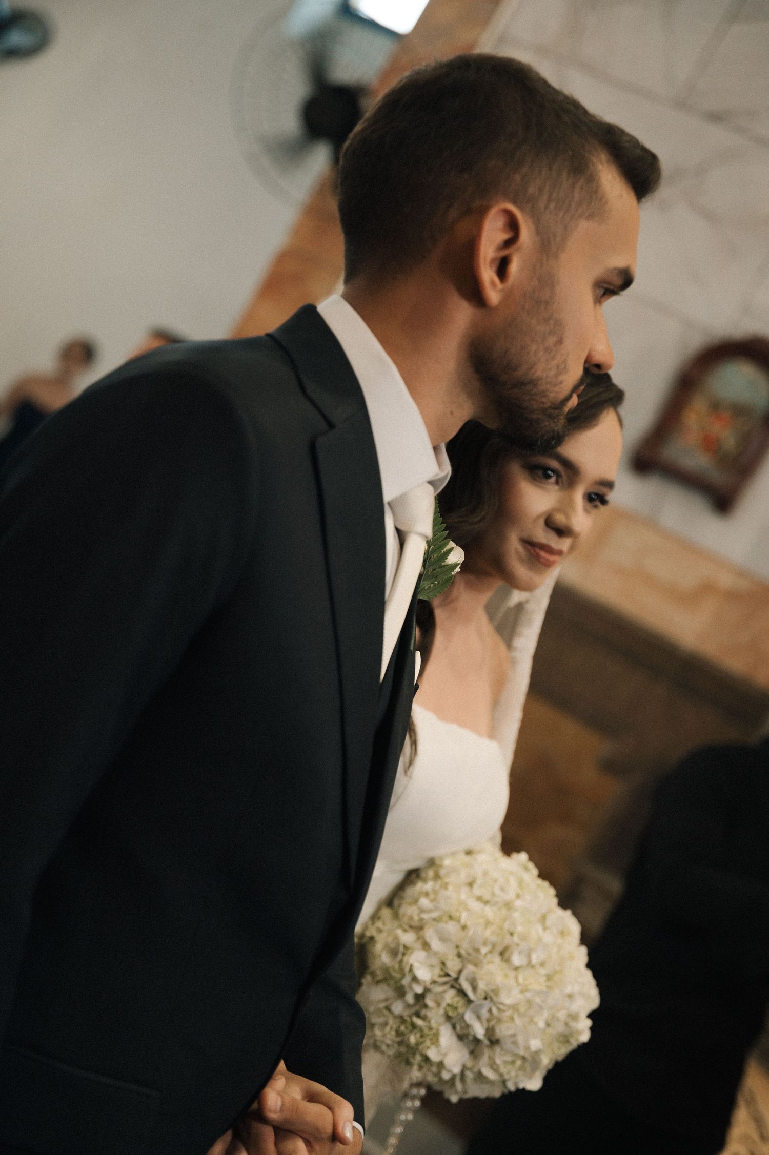 Bride holding white hydrangea bouquet stands beside groom in black suit at church wedding ceremony.