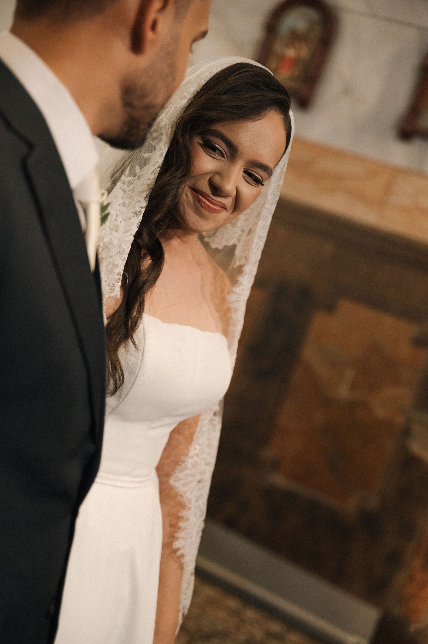 A smiling bride in a white dress and lace veil stands with her groom during a church wedding ceremony.