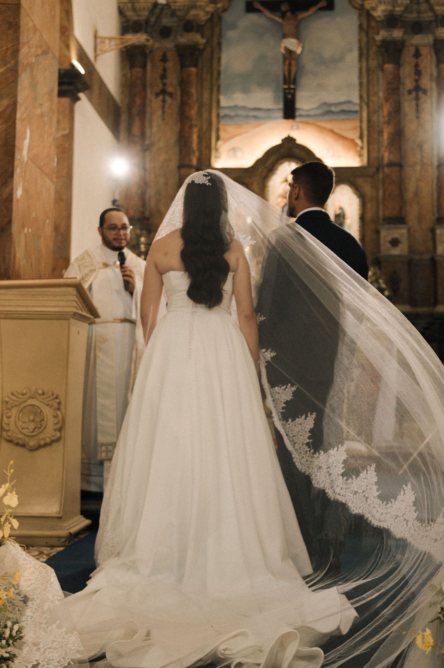 A bride and groom stand before a priest during a Catholic church wedding ceremony, the bride wearing a flowing white gown and lace veil.