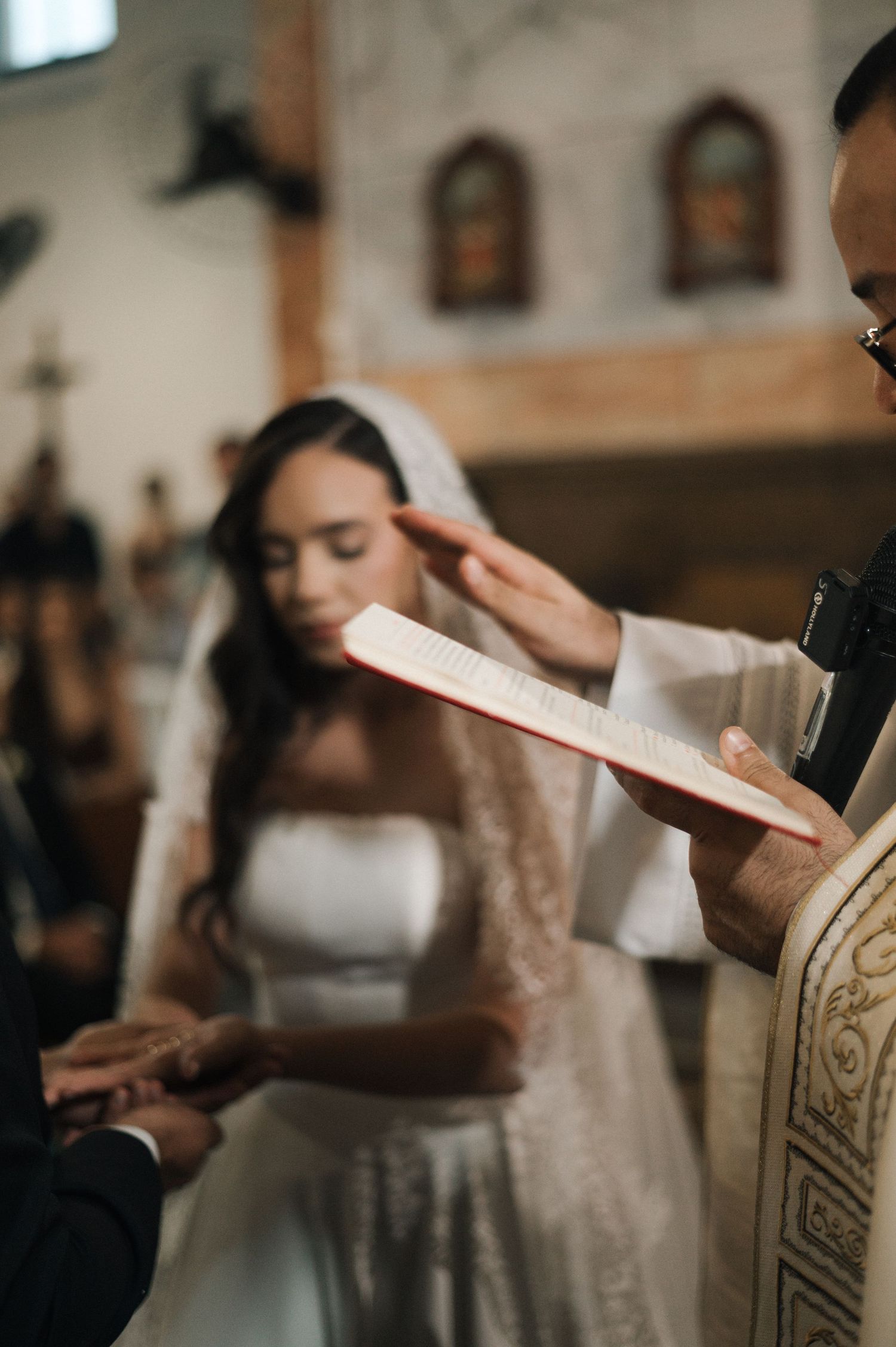 Bride in white lace veil stands at church altar as priest reads from Bible during traditional wedding ceremony.