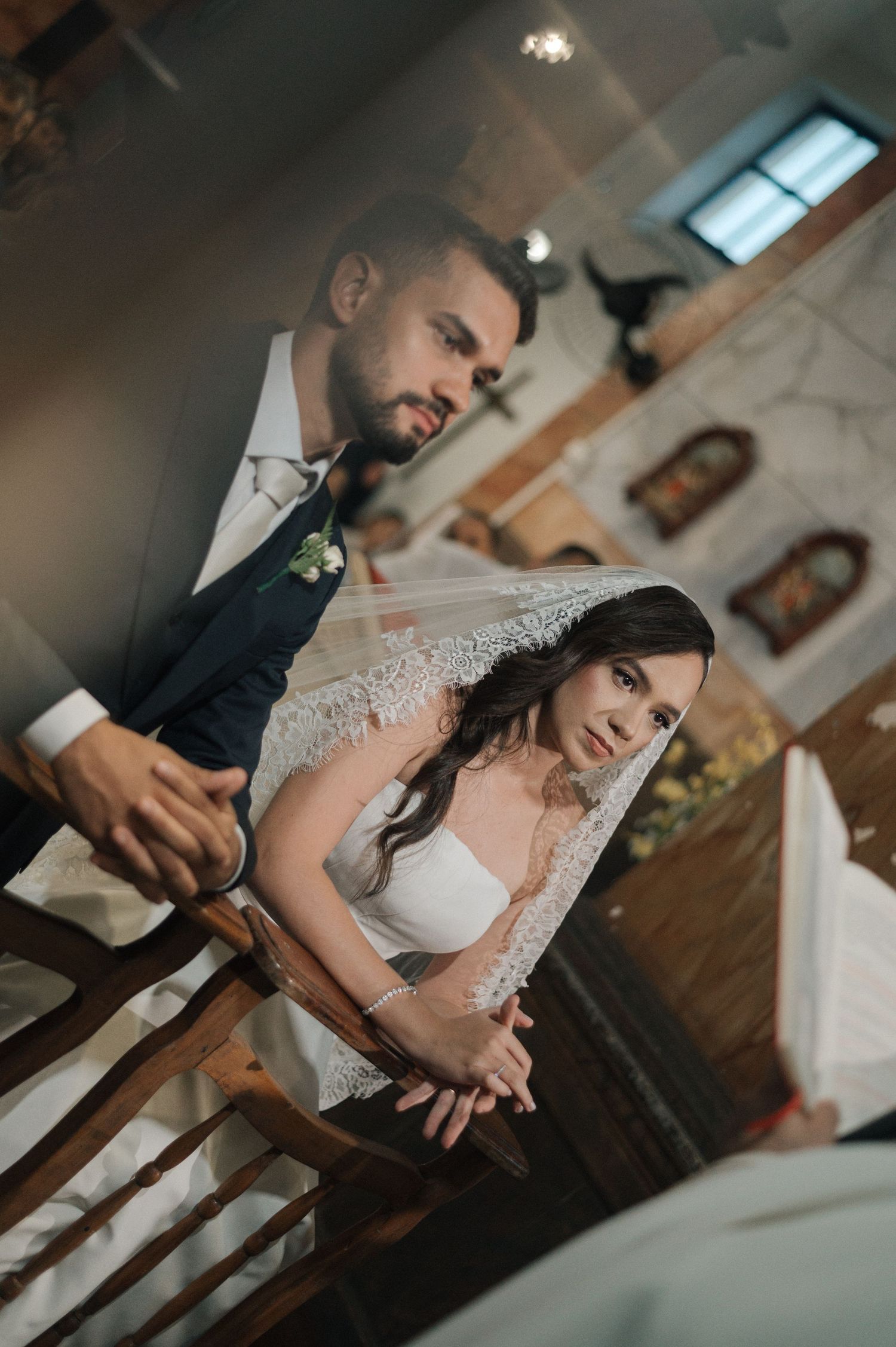 A bride in a white gown and lace veil kneels at a church altar beside her groom in a dark suit during their wedding ceremony.