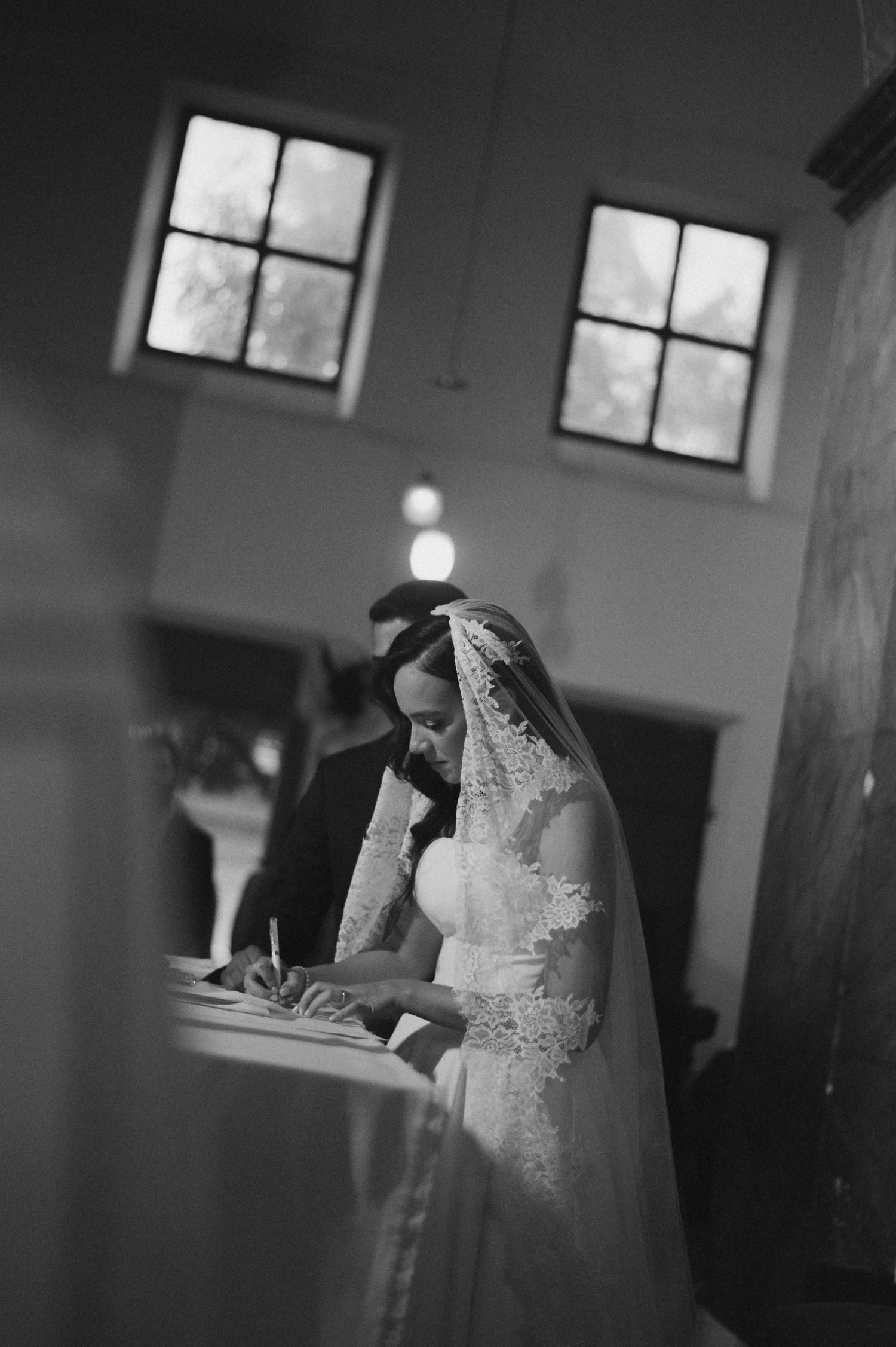 Bride in lace veil signs wedding register beside groom in black suit inside a church.