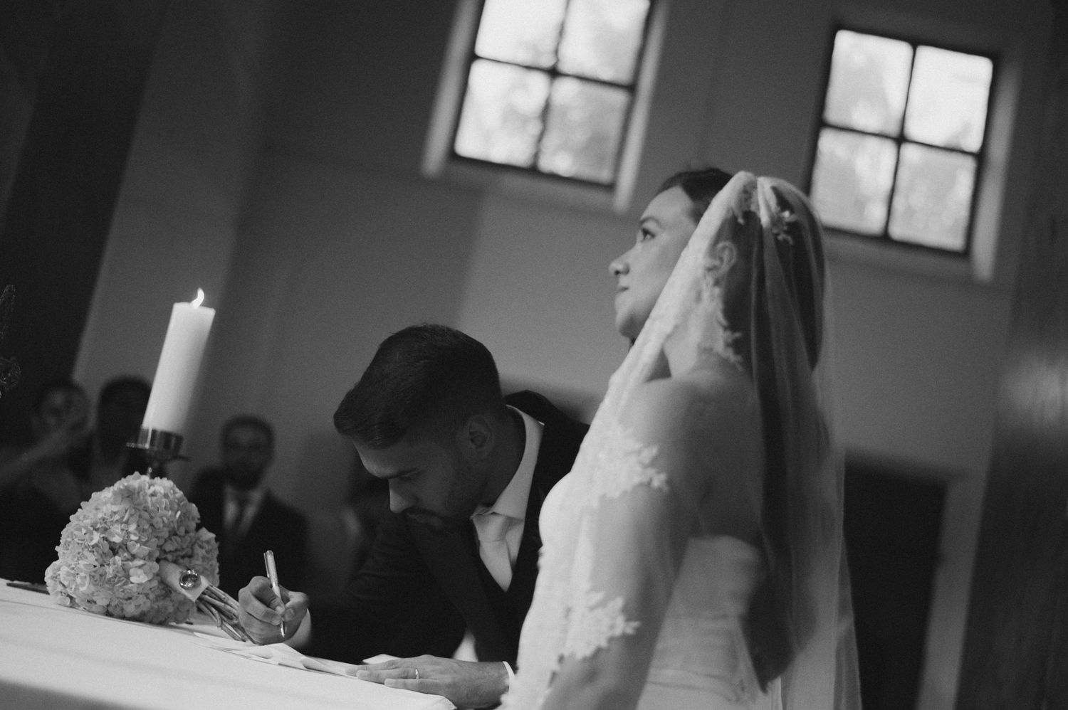 Black and white wedding photo of bride and groom signing marriage certificate at church altar with candle and bouquet.