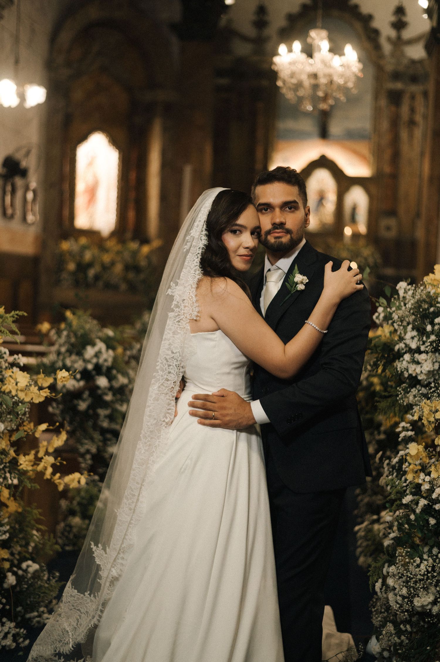 Bride and groom embrace in elegant church adorned with white and yellow floral arrangements during wedding ceremony.
