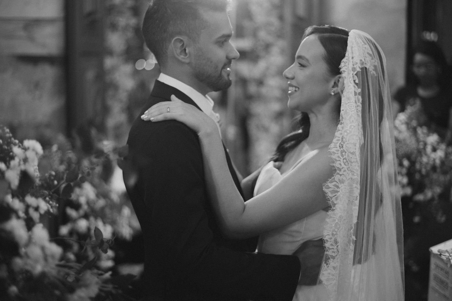 Black and white wedding photo of a smiling bride in lace veil and groom in tuxedo embracing at floral altar.