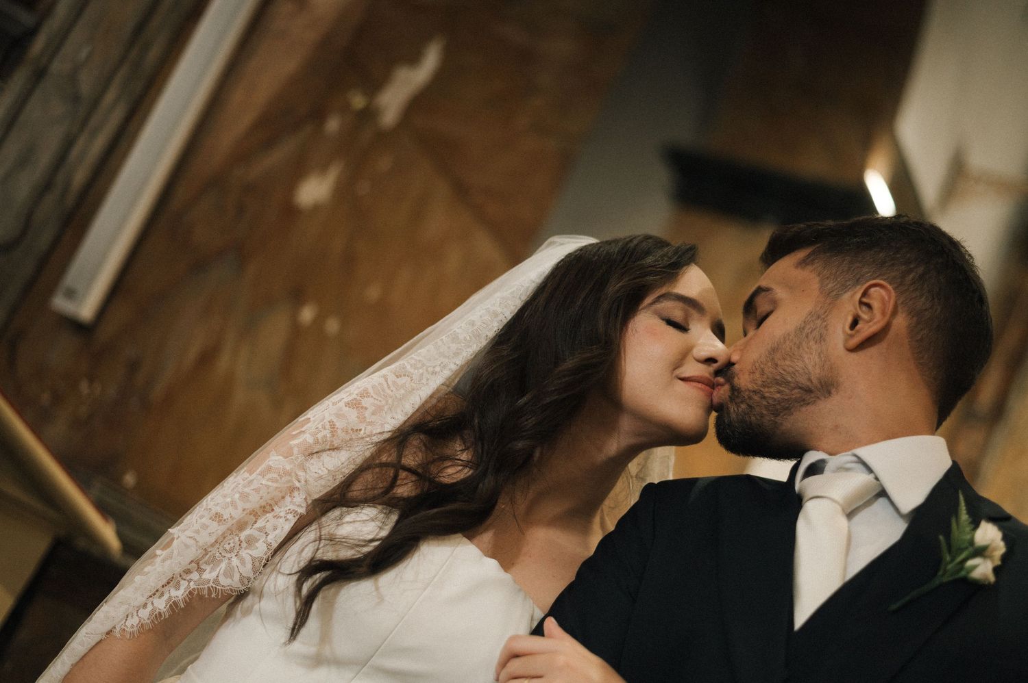 A bride and groom share a romantic kiss at their wedding, with the bride wearing a lace veil and white gown.