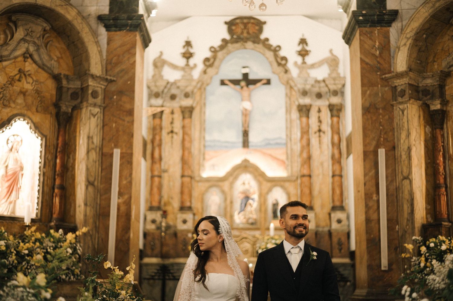 Bride and groom stand at ornate Catholic church altar with crucifix and marble decor during wedding ceremony.