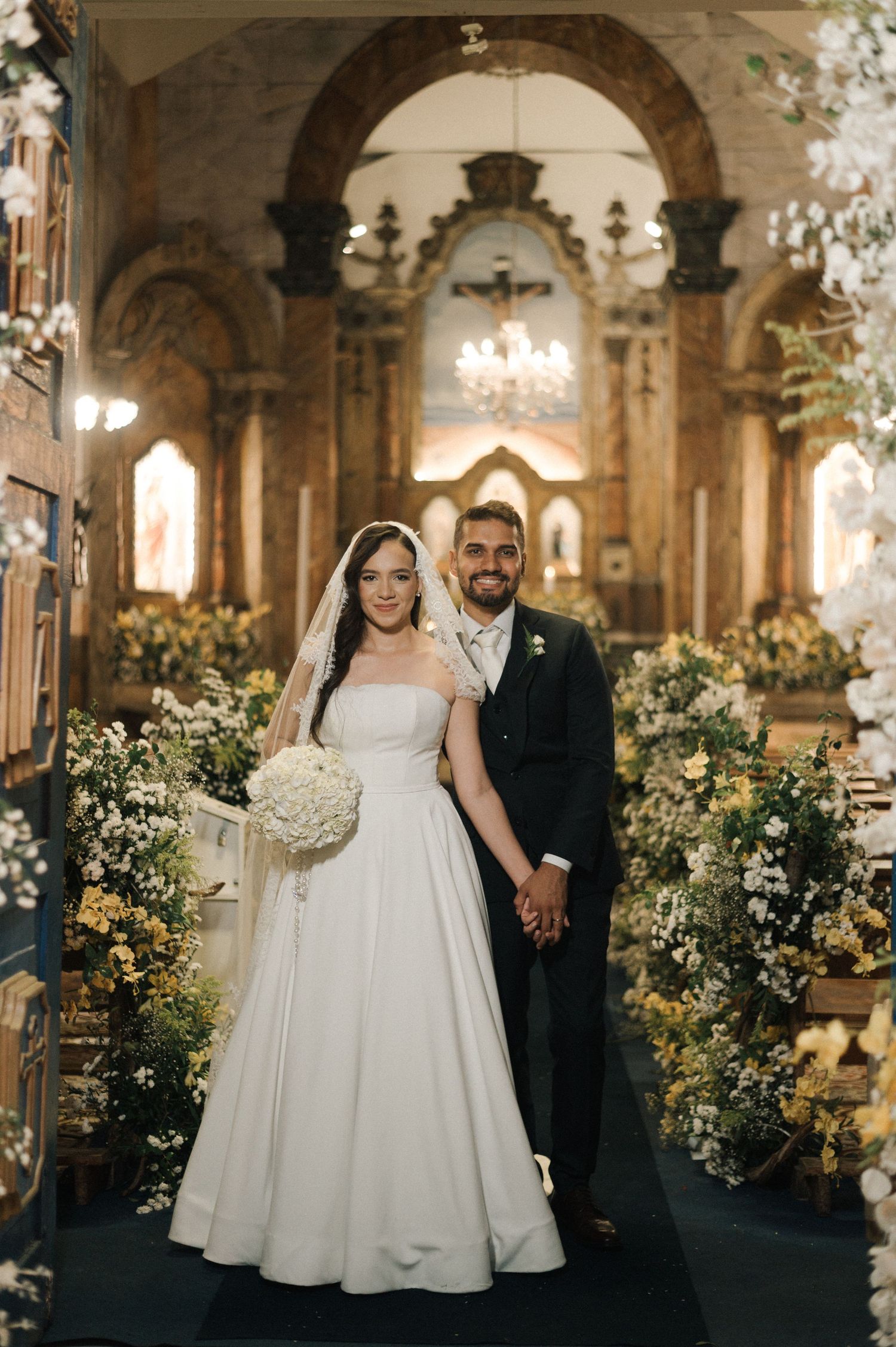 Bride and groom pose together in an ornate church adorned with white floral arrangements after their wedding ceremony.