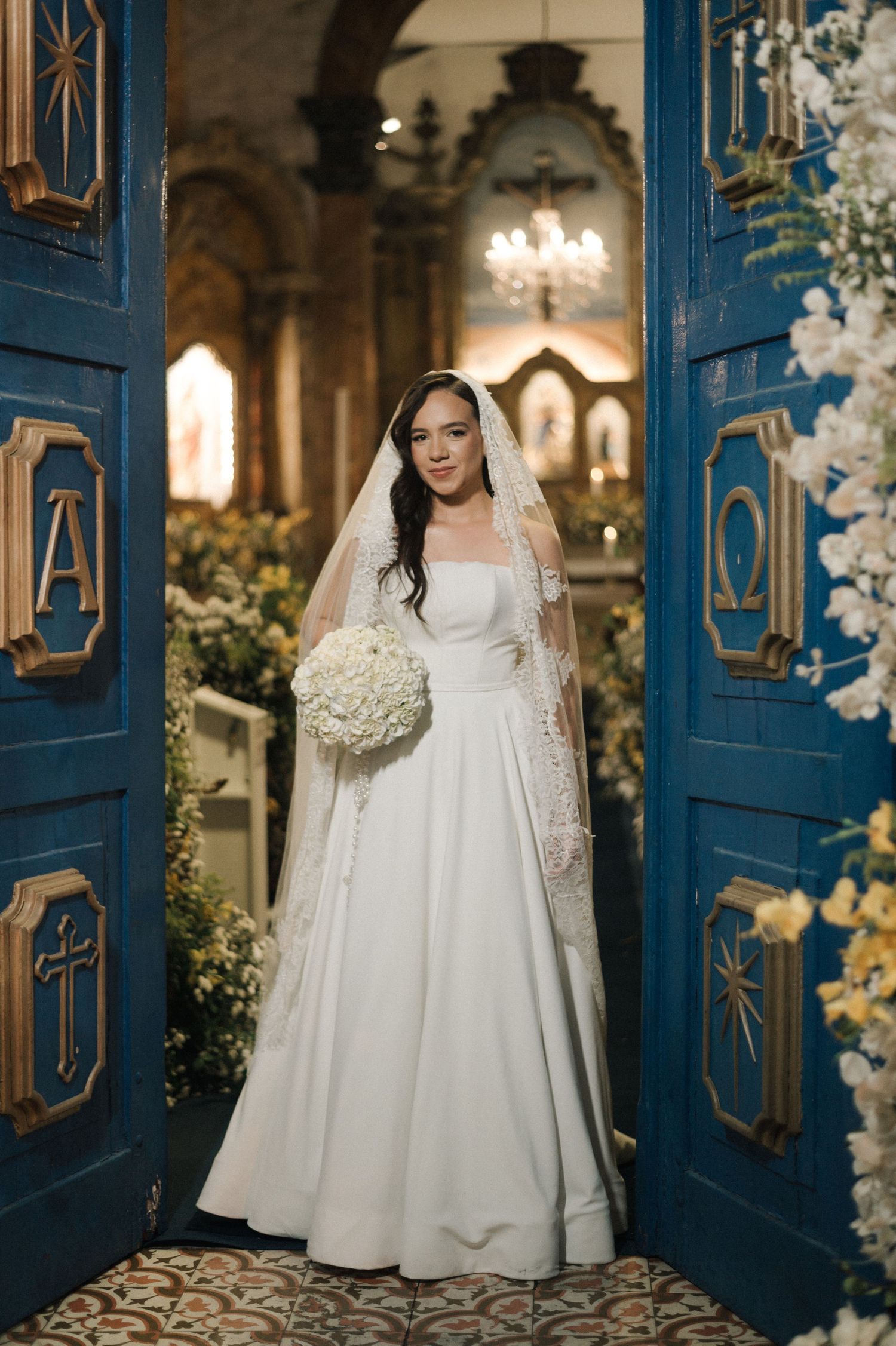 Bride in white gown and lace veil holds bouquet at ornate blue church doors adorned with religious symbols.