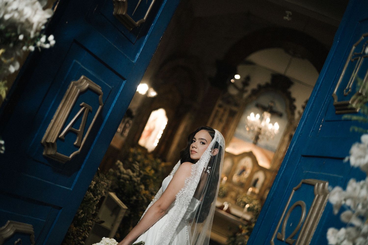 Bride in white lace gown and veil stands before ornate blue doors in an elegant church setting.