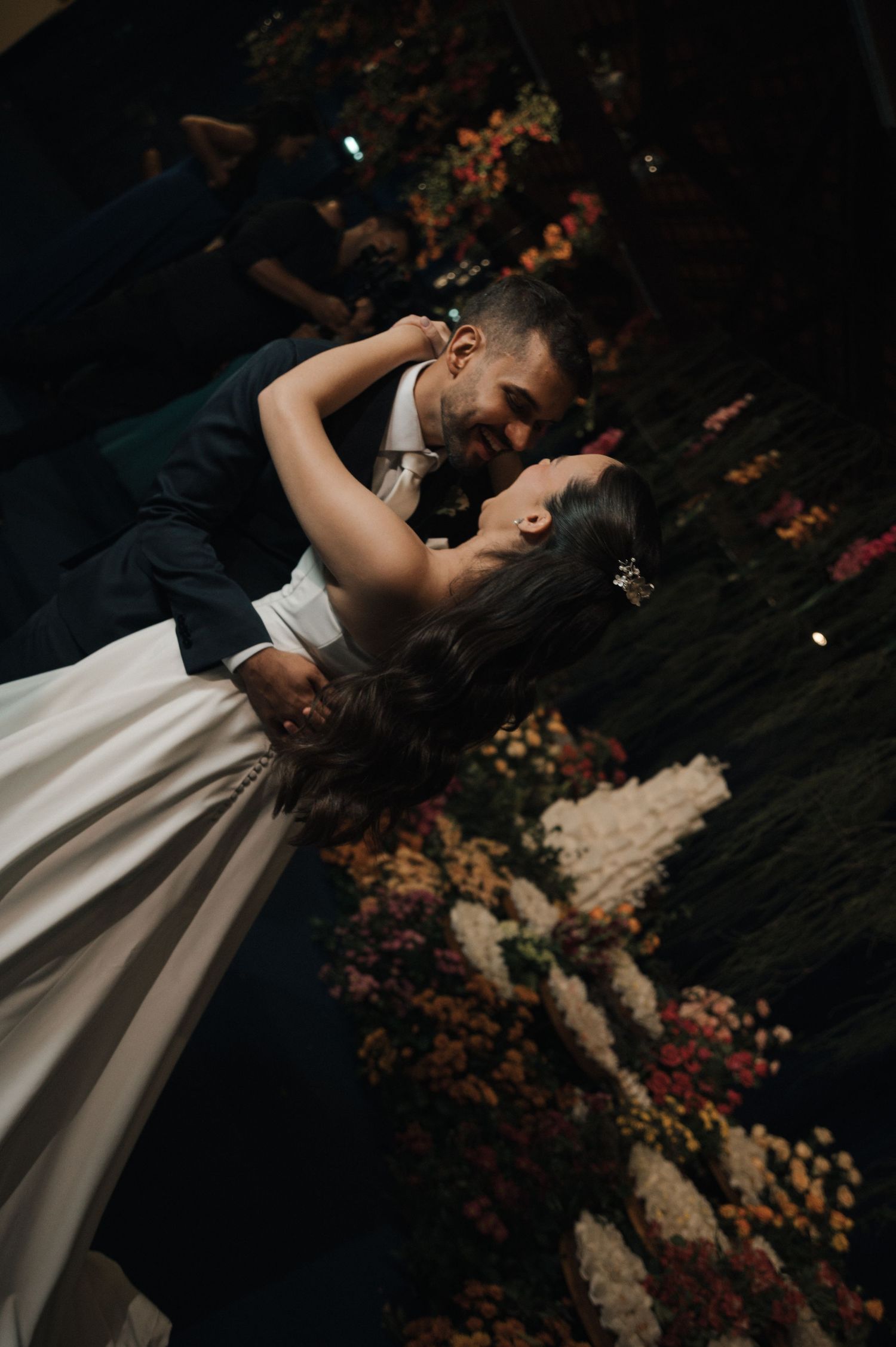 A couple shares a romantic dip kiss on their wedding dance floor surrounded by lush floral arrangements.