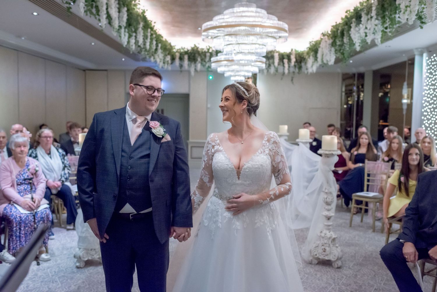 A smiling bride and groom hold hands at the altar during their elegant wedding ceremony with floral ceiling decor.