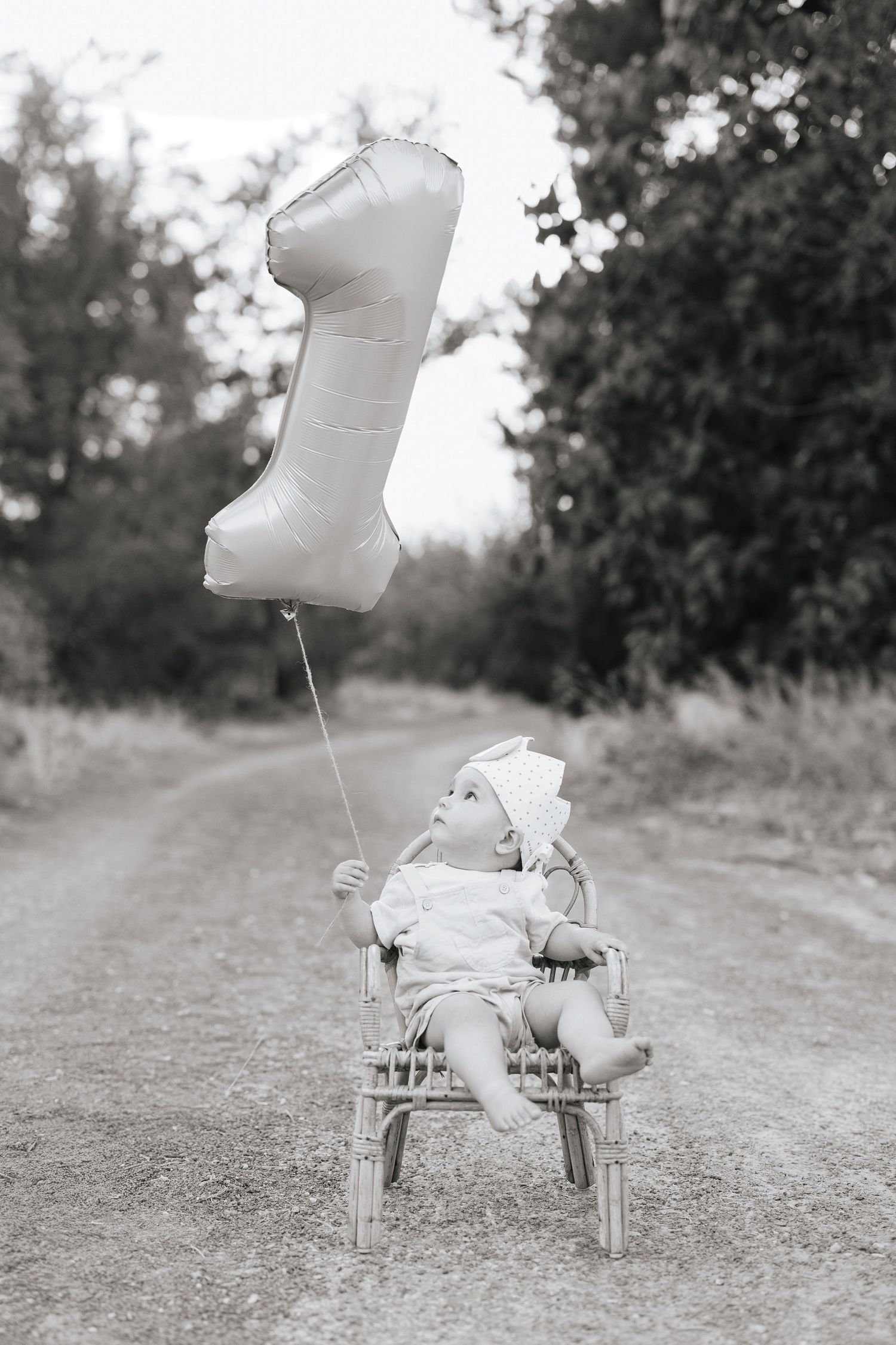 Black and white photo of a baby sitting in a wicker chair holding a number one balloon on a dirt road.