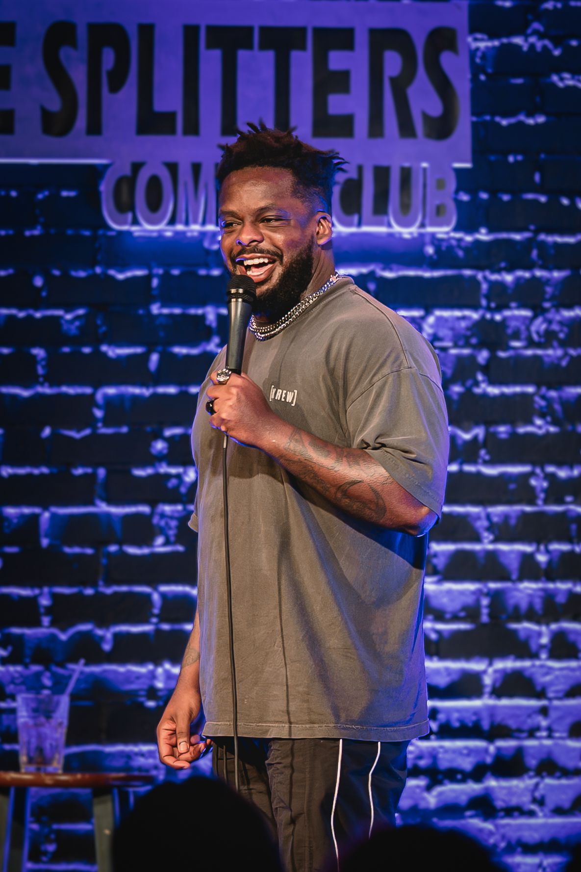 Dedrick Flynn A male stand-up comedian performs on stage at a comedy club, holding a microphone in front of a brick wall backdrop.