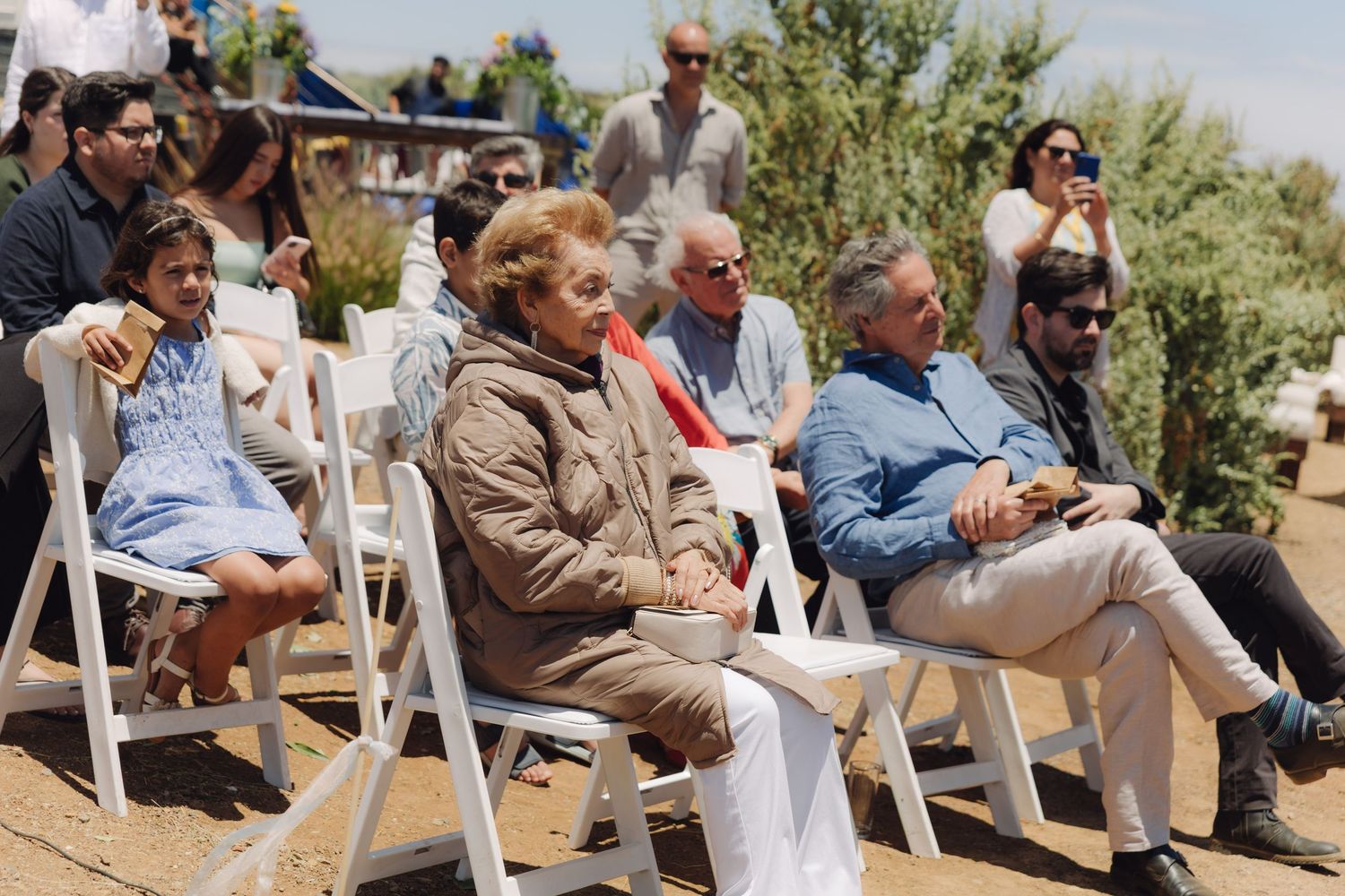 Wedding guests of various ages seated in white chairs at sunny outdoor ceremony.