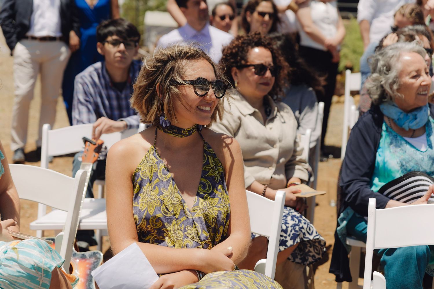 Smiling woman in floral dress with sunglasses enjoying outdoor wedding ceremony as a guest.
