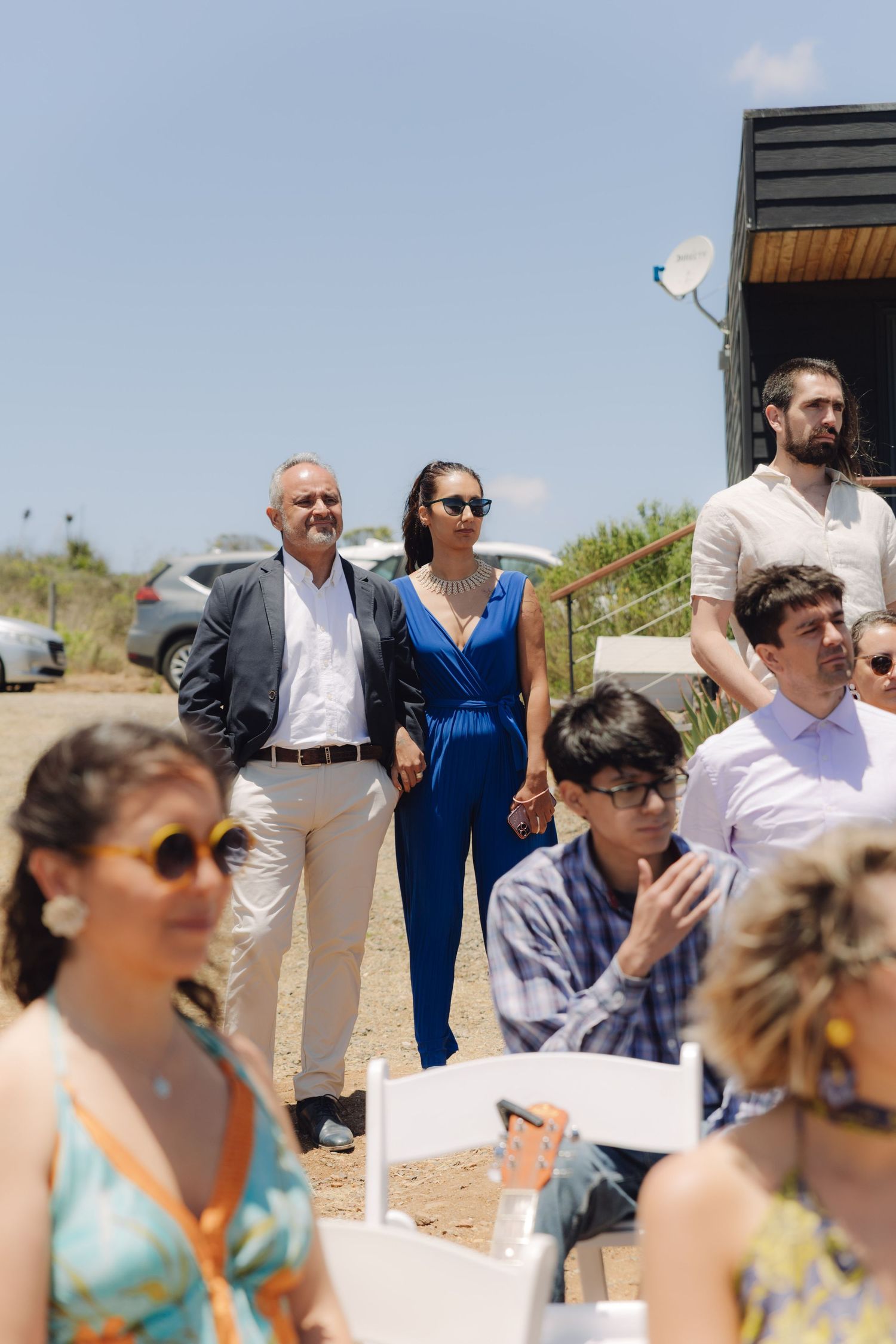 Wedding guests standing and seated outdoors in bright sunlight watching the ceremony.