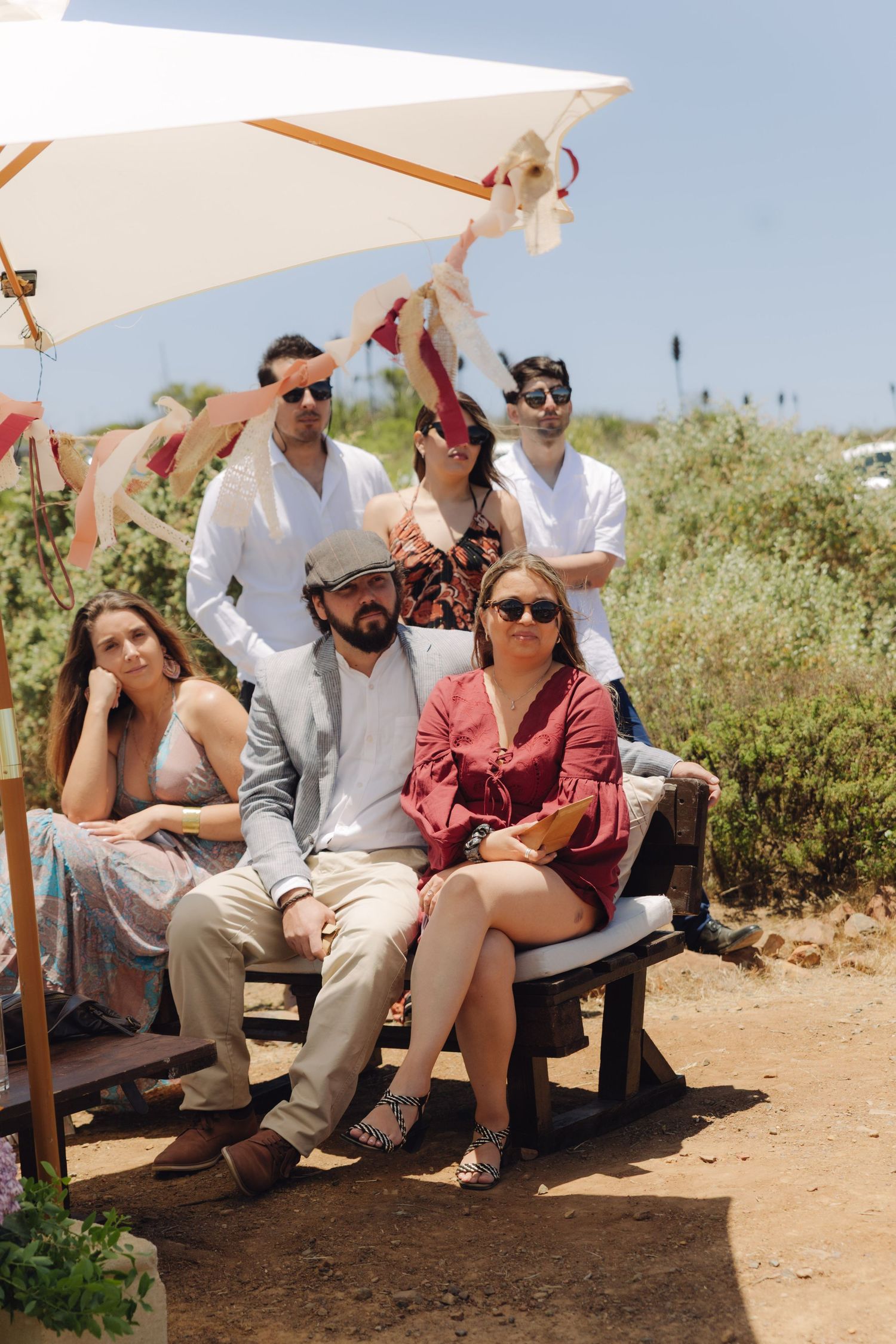 Group of wedding guests relaxing together under a patio umbrella at outdoor celebration.