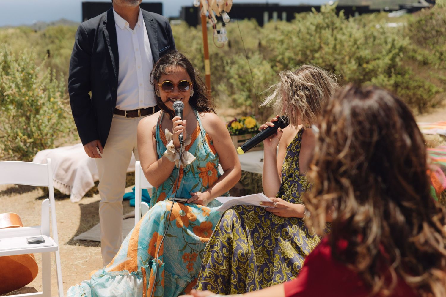 Woman in floral dress speaking into microphone at outdoor wedding ceremony.