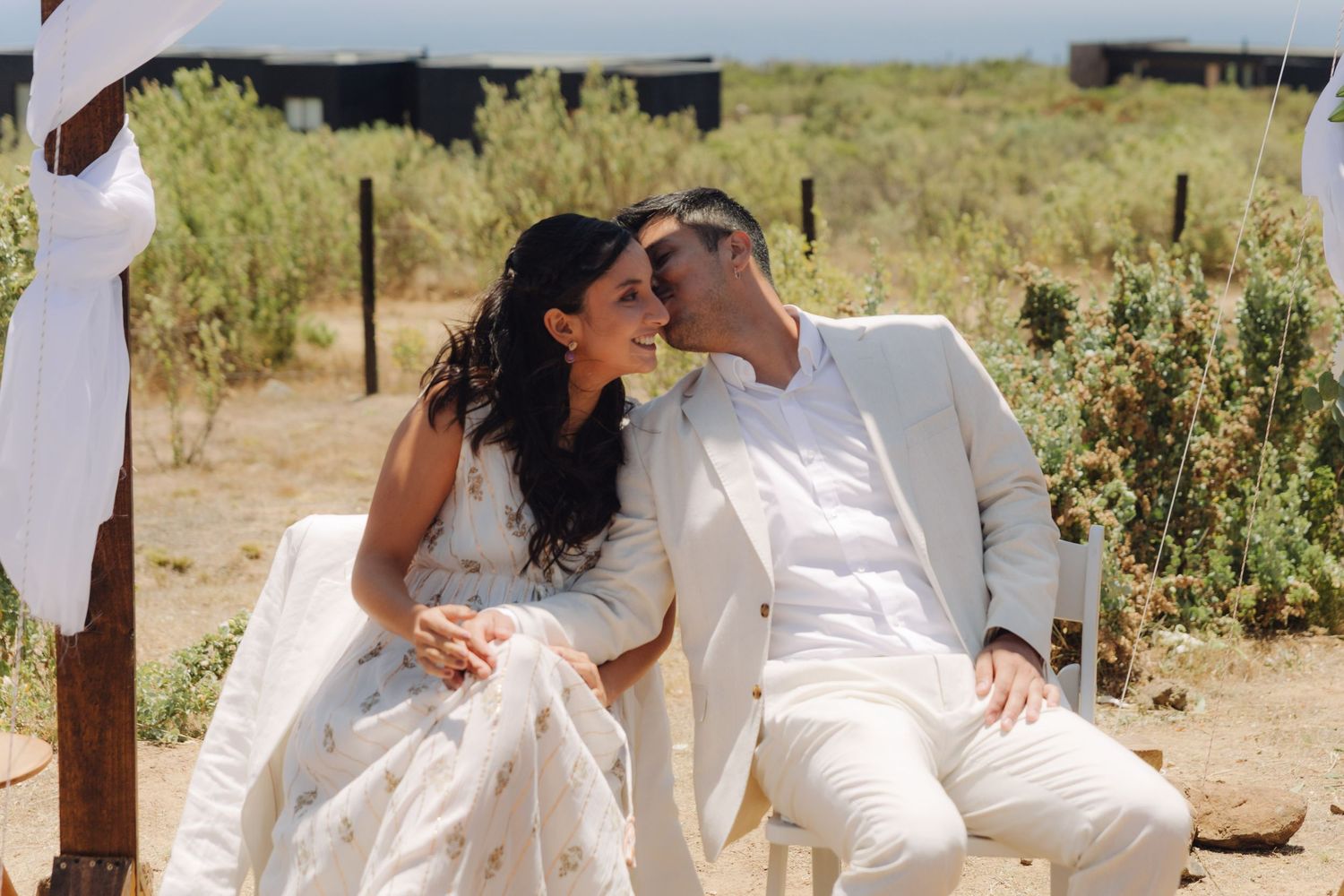 Bride and groom in white attire sharing intimate moment at outdoor wedding.