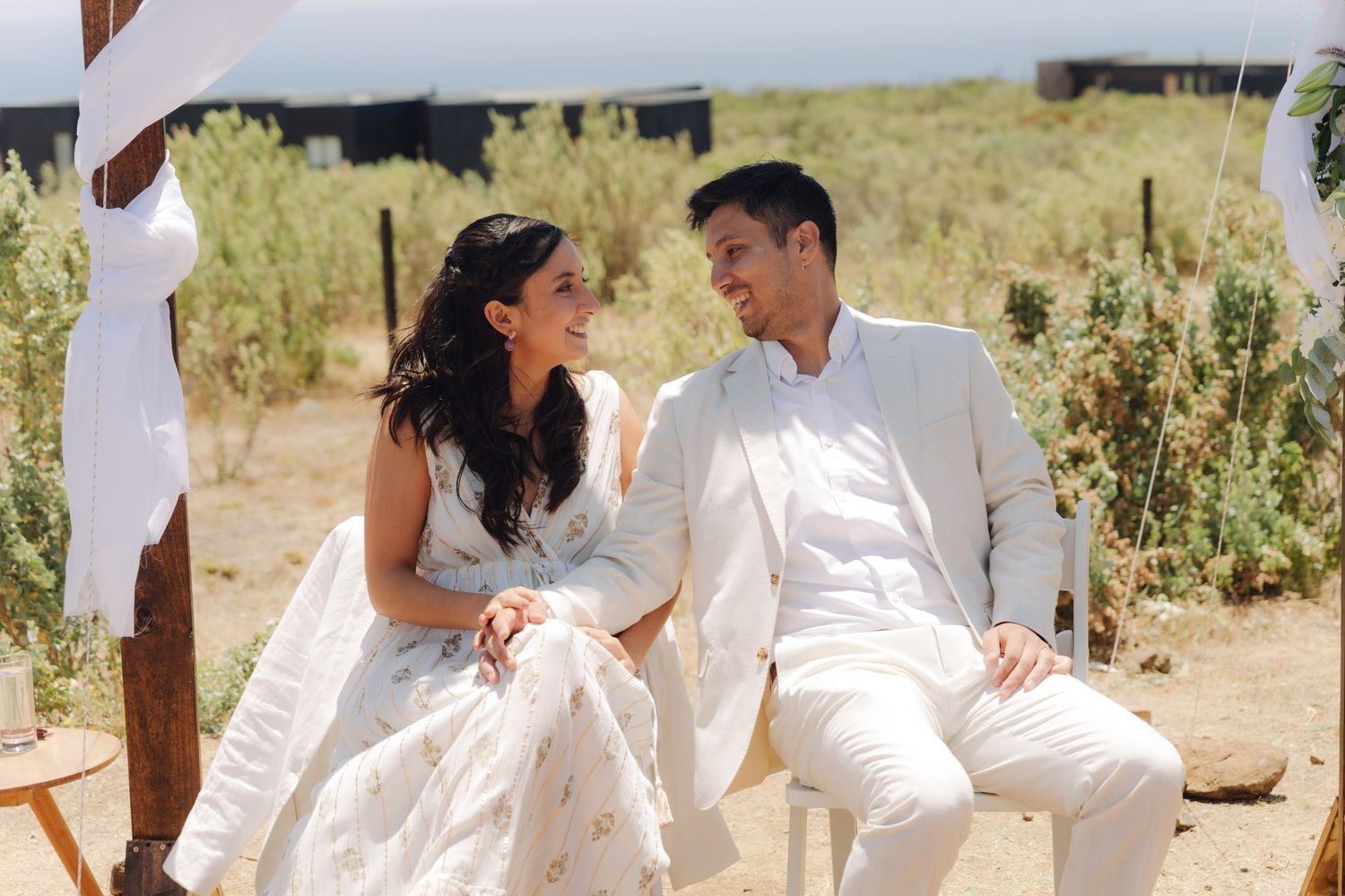 Bride in white gown and groom in white suit exchanging smiles at wedding.