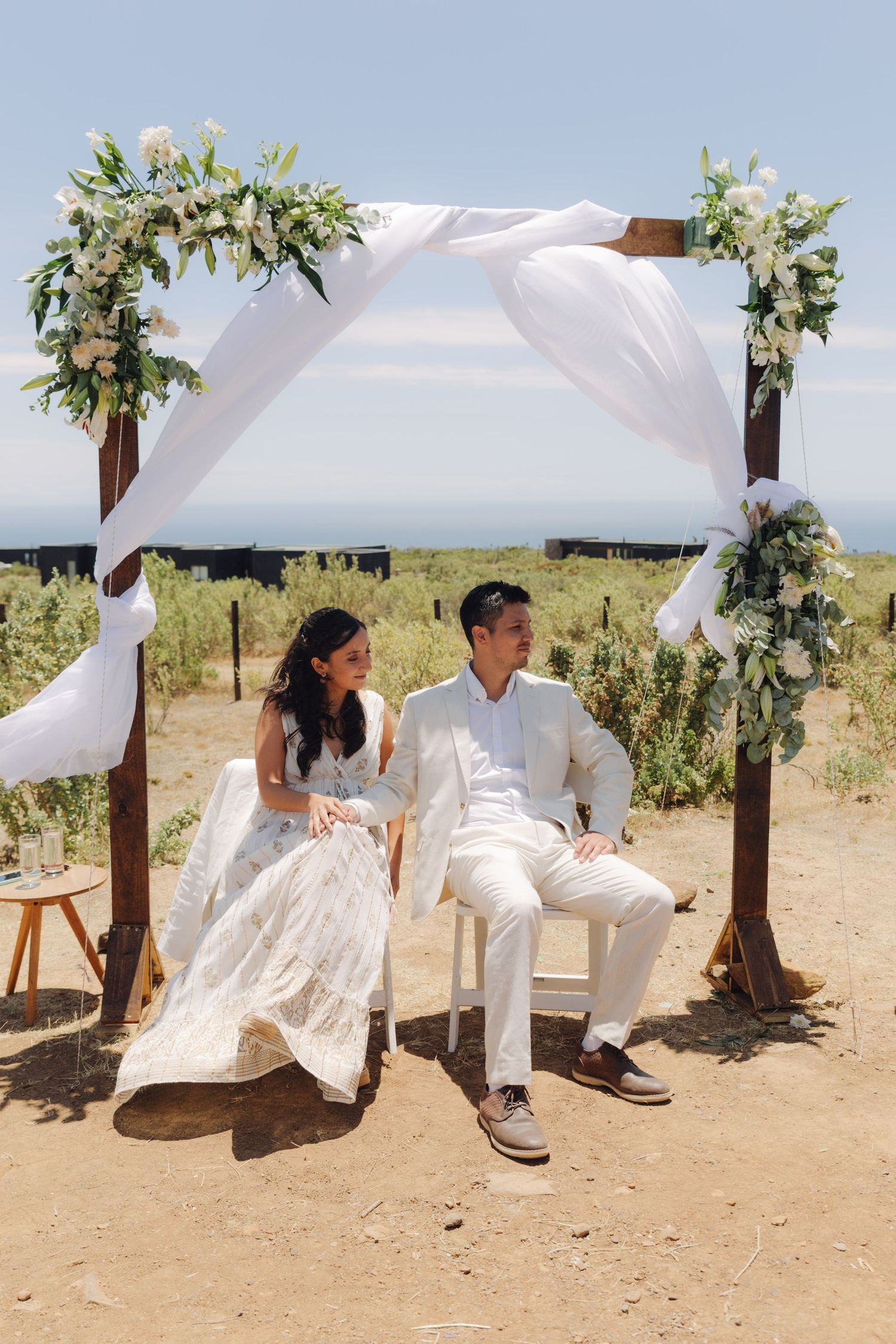 Bride and groom seated under floral wooden arch at outdoor beach wedding.