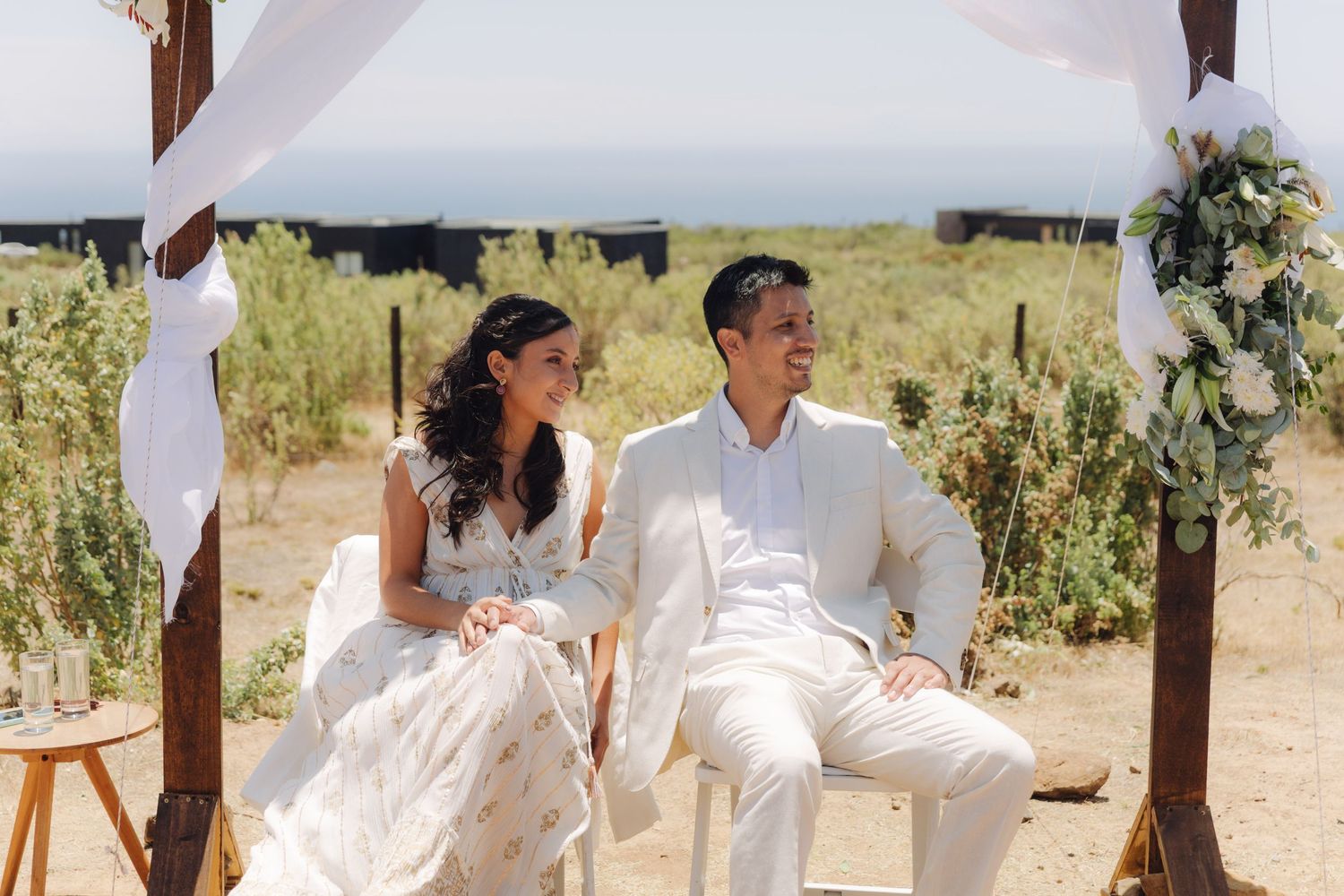 Smiling bride and groom seated under decorated wooden arch at outdoor wedding.