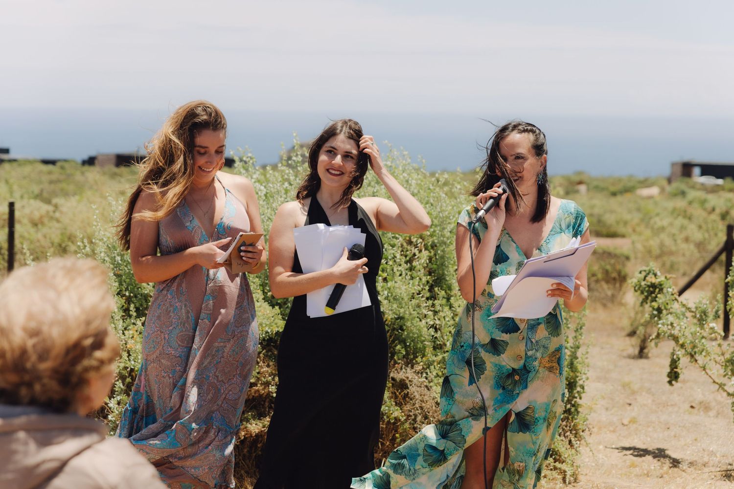 Three women smiling and holding papers at outdoor wedding ceremony.