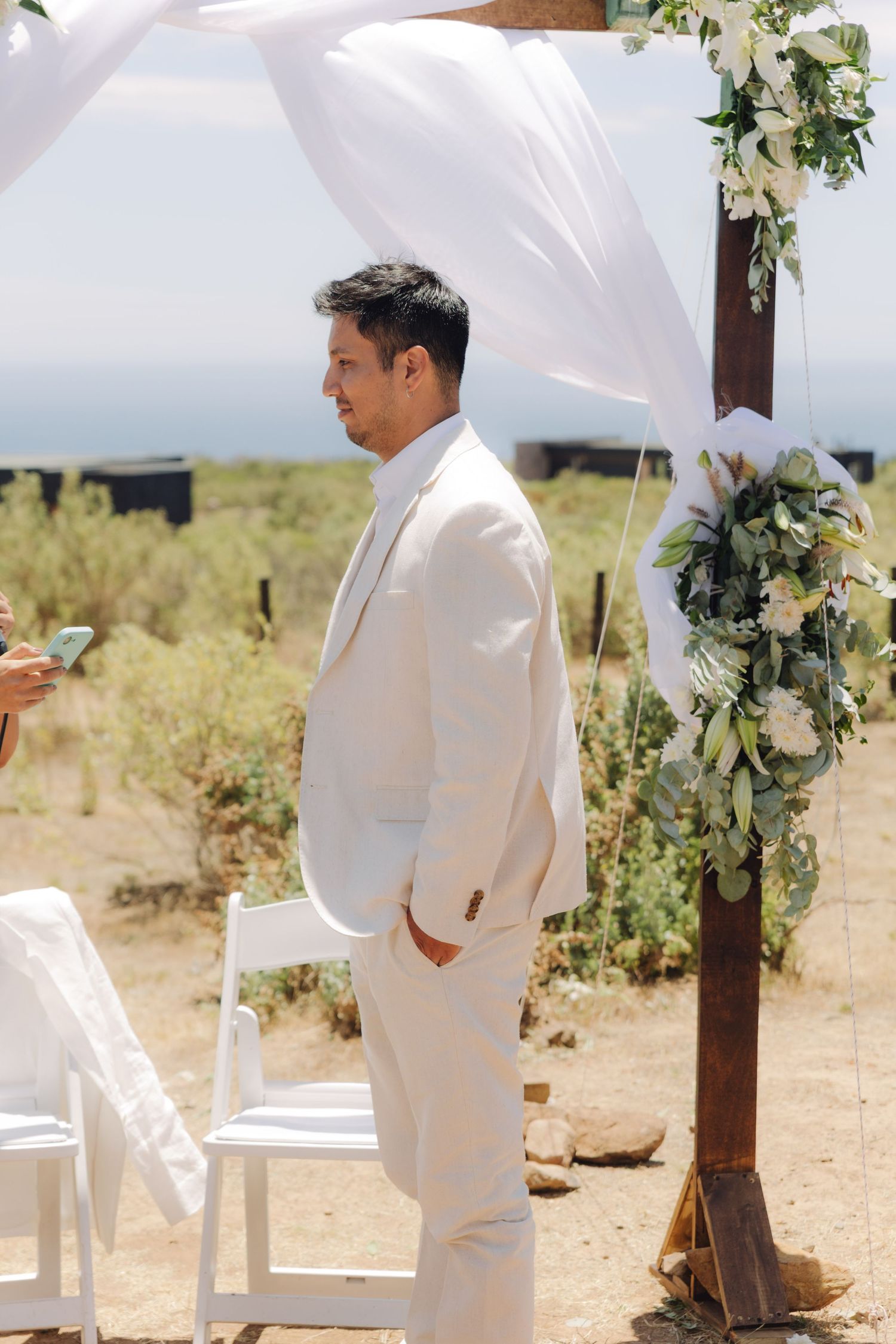 Groom in cream suit stands at outdoor wedding altar decorated with white draping and floral arrangements.