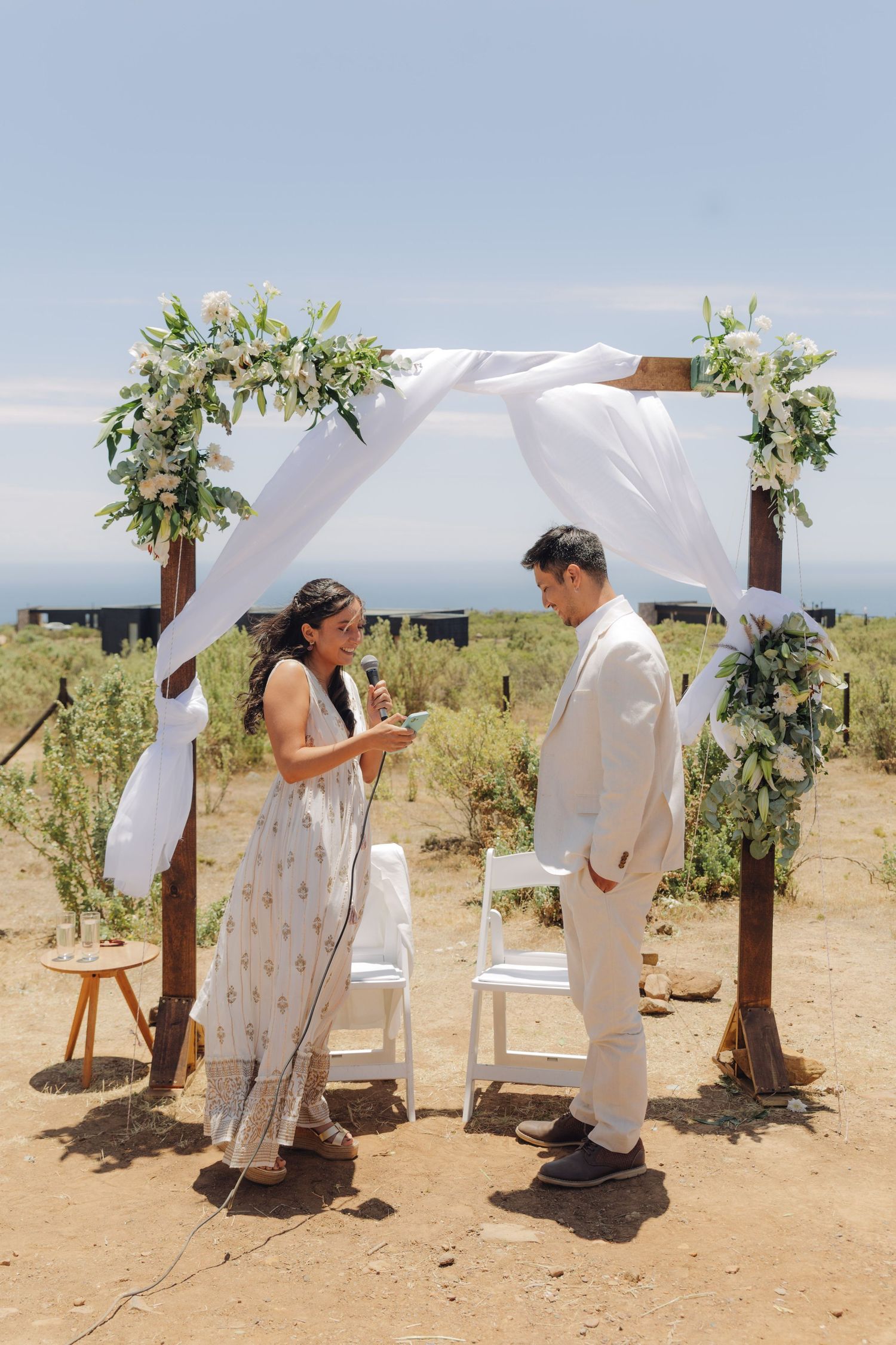 Bride and groom exchange vows under a rustic wooden arch with white draping and greenery at outdoor wedding.
