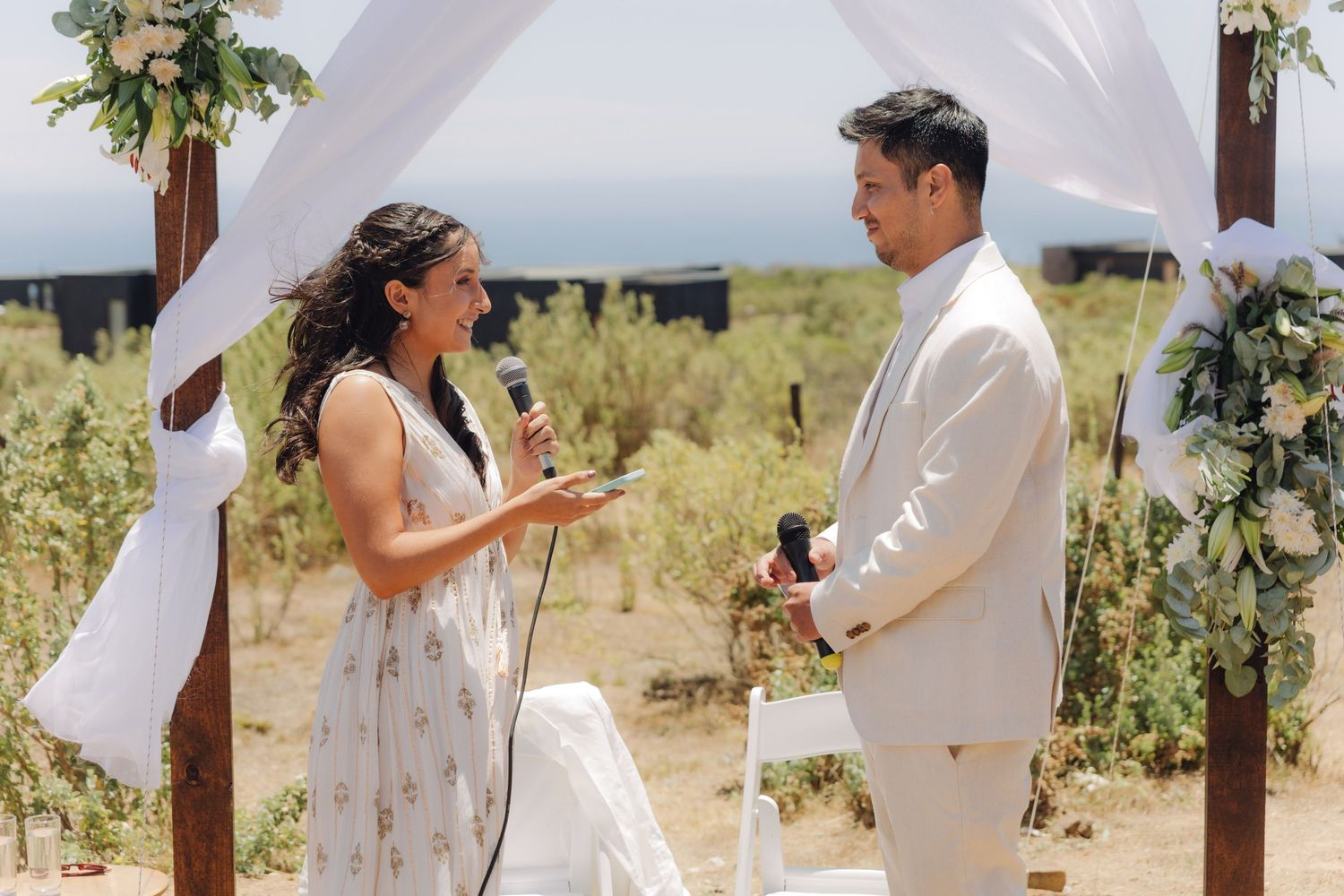 Bride reads wedding vows into microphone while groom listens attentively at outdoor desert ceremony.