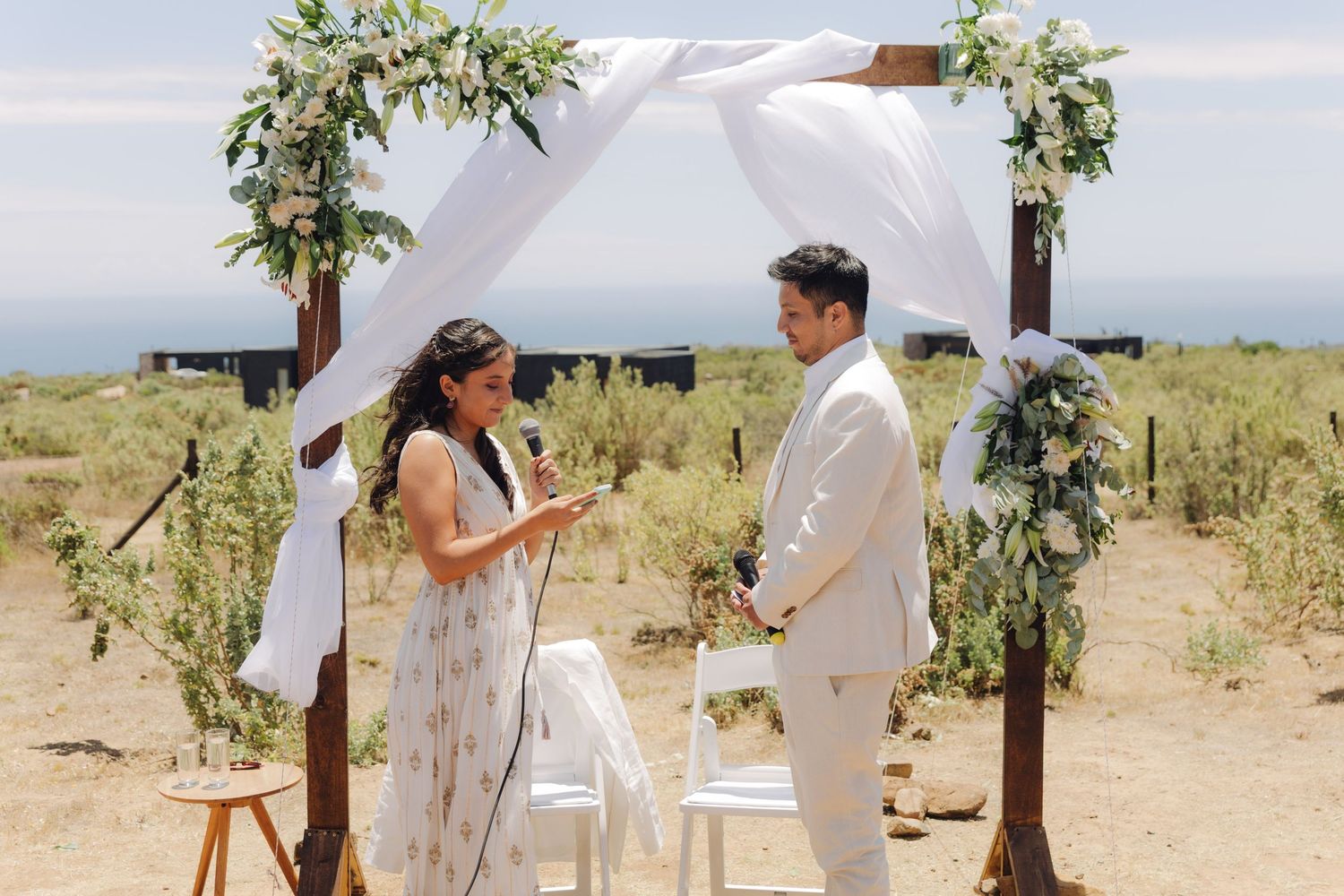 Couple stands facing each other during outdoor wedding ceremony under floral arch with ocean view backdrop.